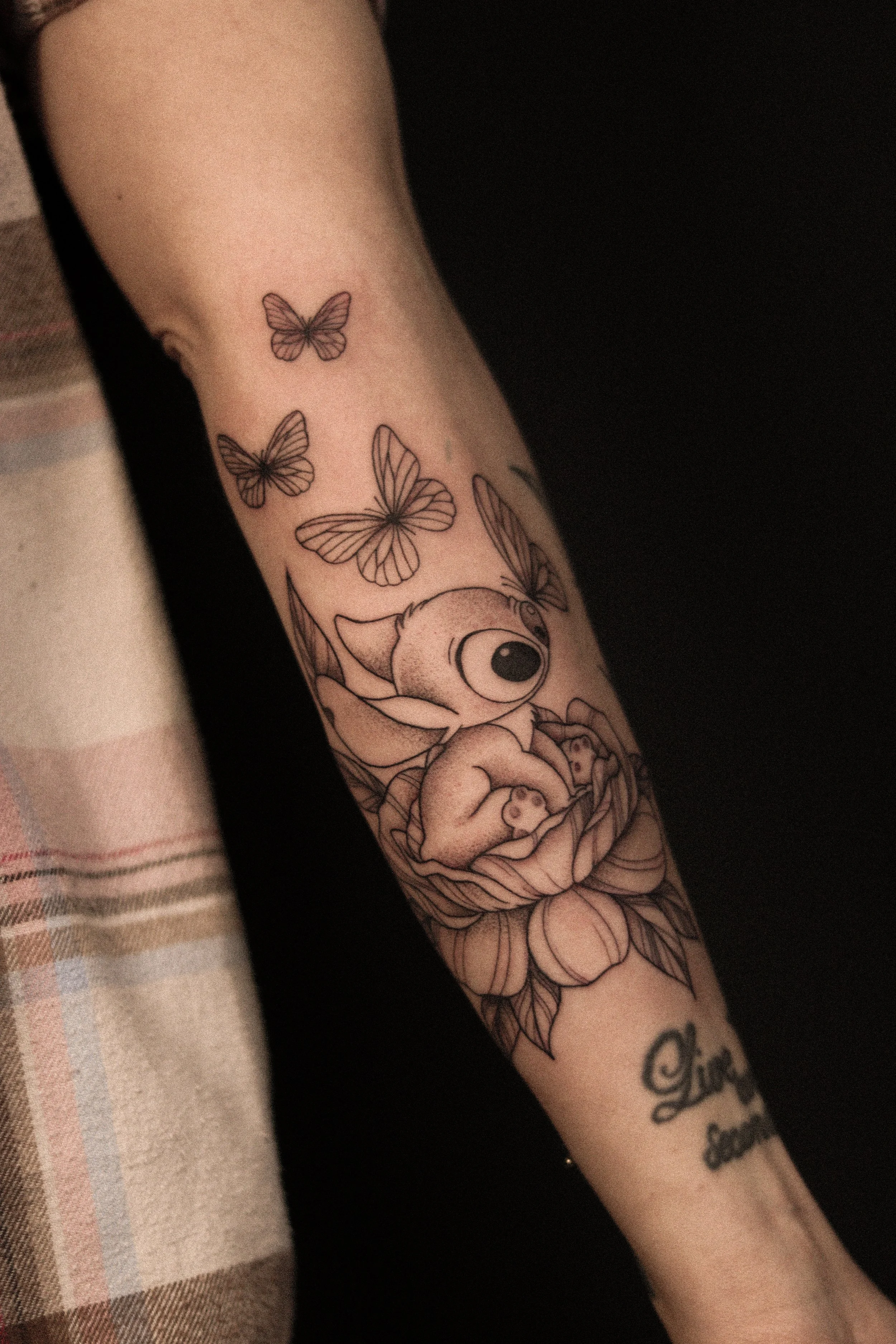 Tattoo op de onderarm van een dier dat in een bloem ligt, omlijst door vliegende vlinders en een tekst onder de bloem.