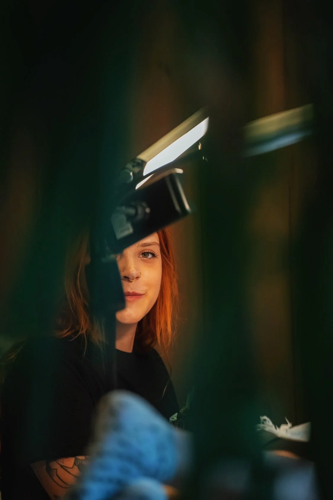 Portret van een jonge vrouw met rood haar en een neusring, die door een papierknipkunstwerk kijkt. De foto is genomen door een kleine opening in het kunstwerk, die haar gedeeltelijk bedekt.