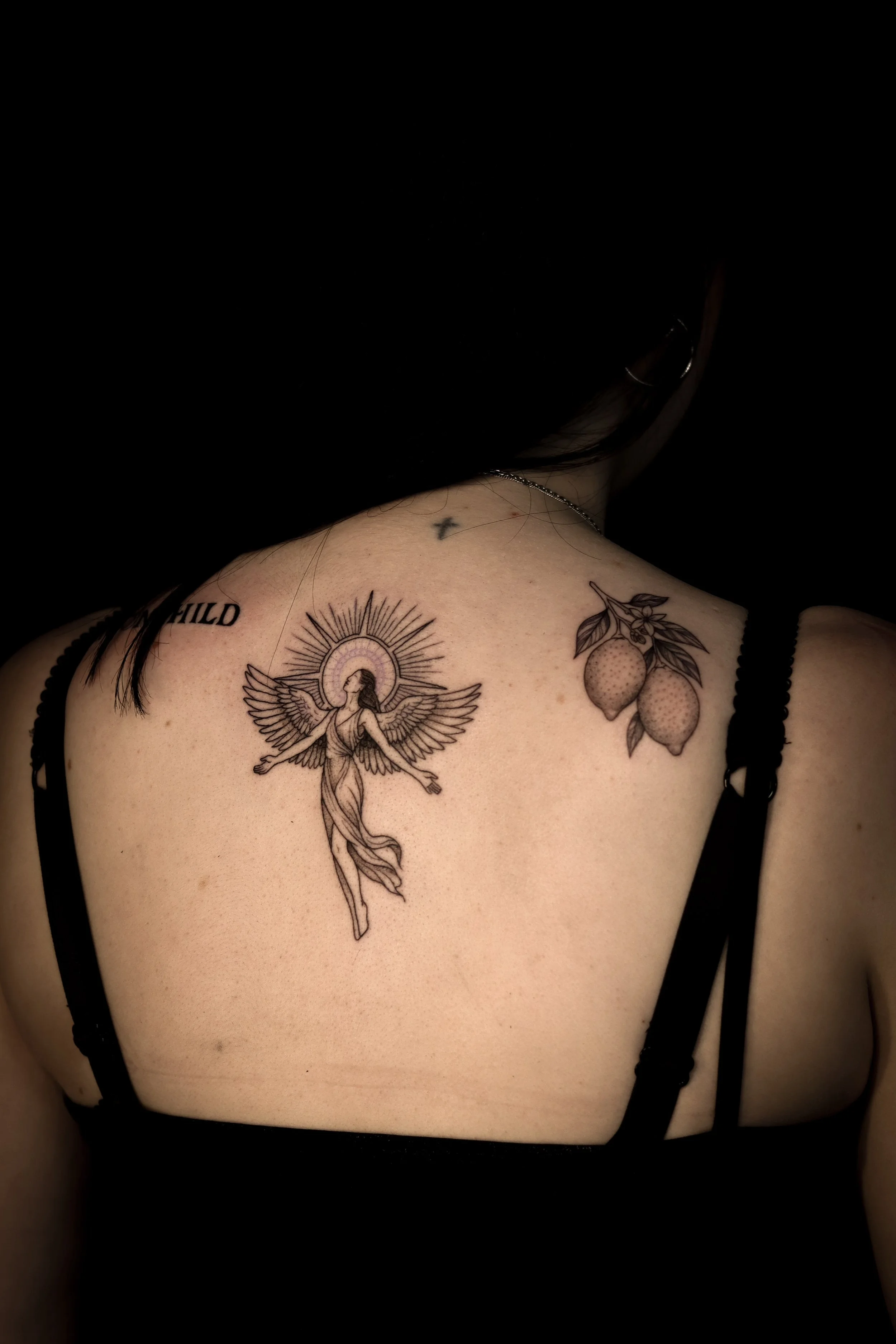 Tattoo van een engel met uitgestrekte armen en vleugels, omringd door een halo, op de schouder van een persoon met een zwarte top.