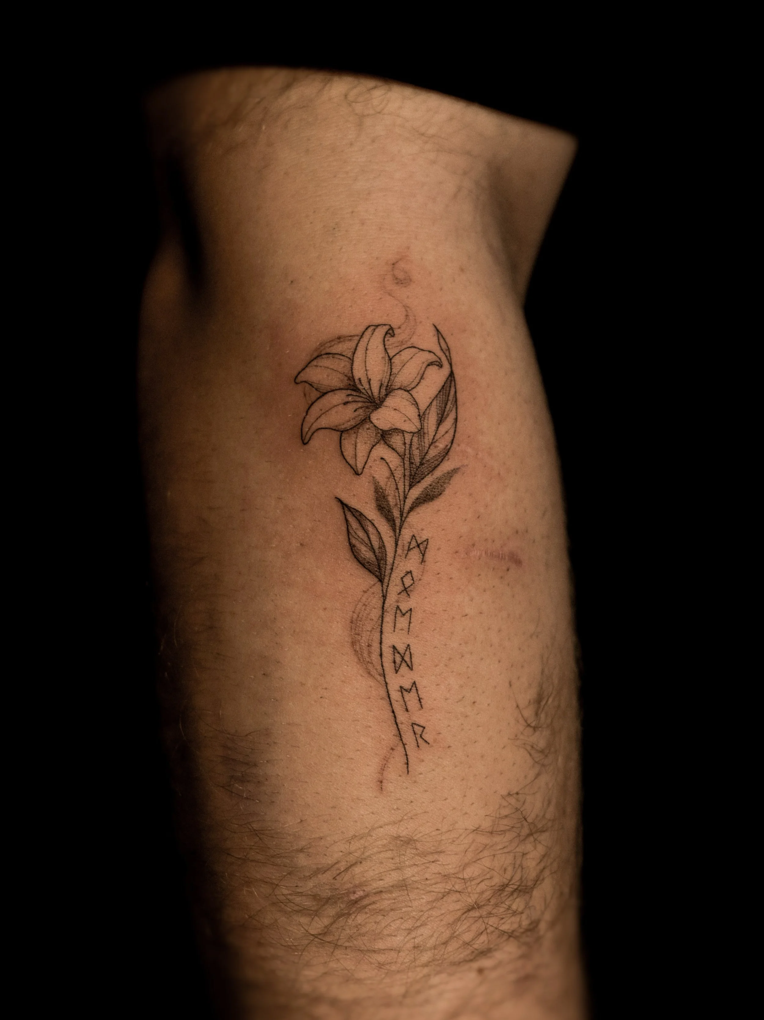 Tattoo van een lis, met bladeren en een golfvorm, en het woord 'MAMOR' onderaan op de binnenkant van de onderarm.