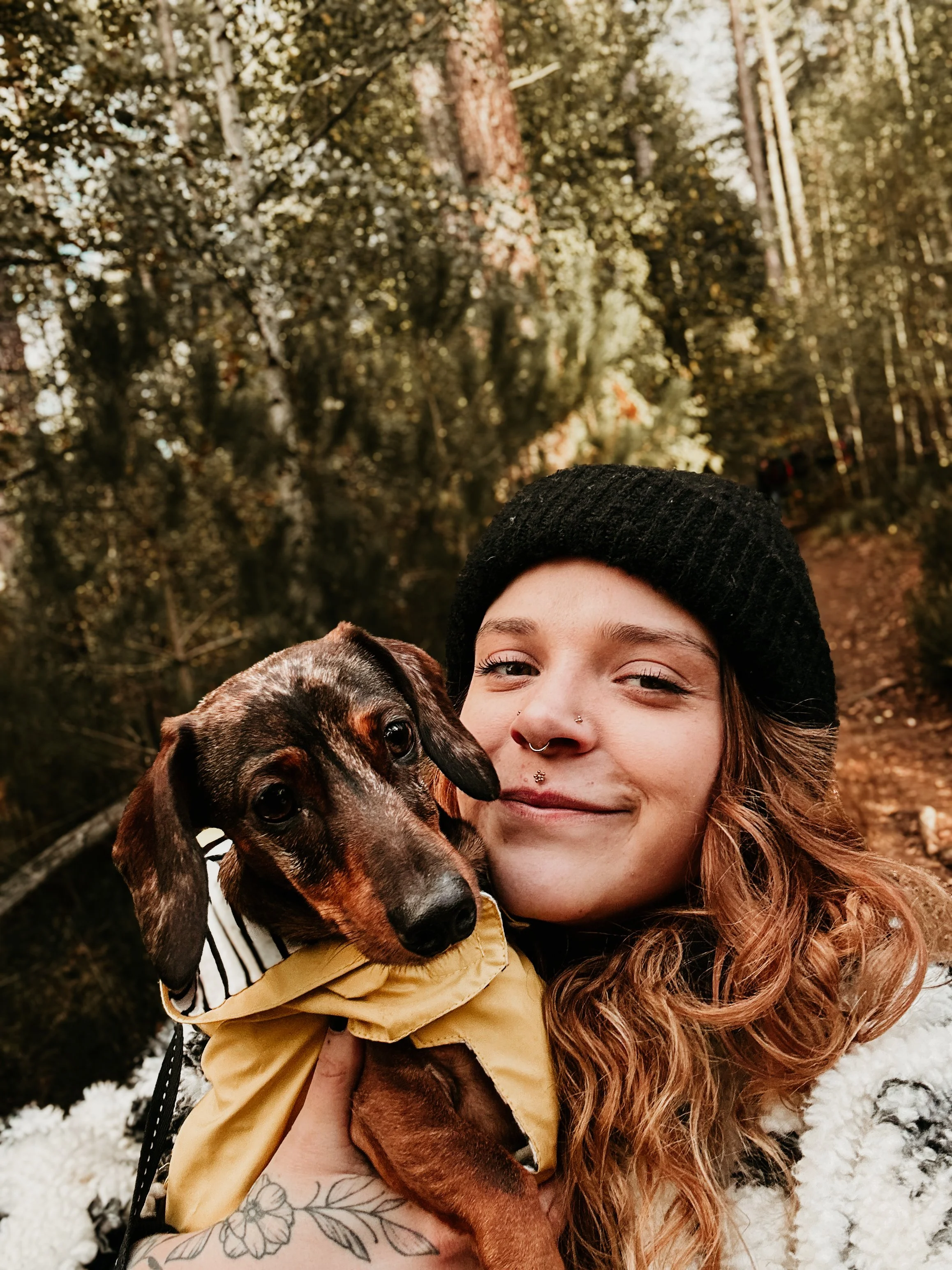 Vrouw met rood haar houdt een kleine bruine teckel in een bos, beiden kijken naar de camera.