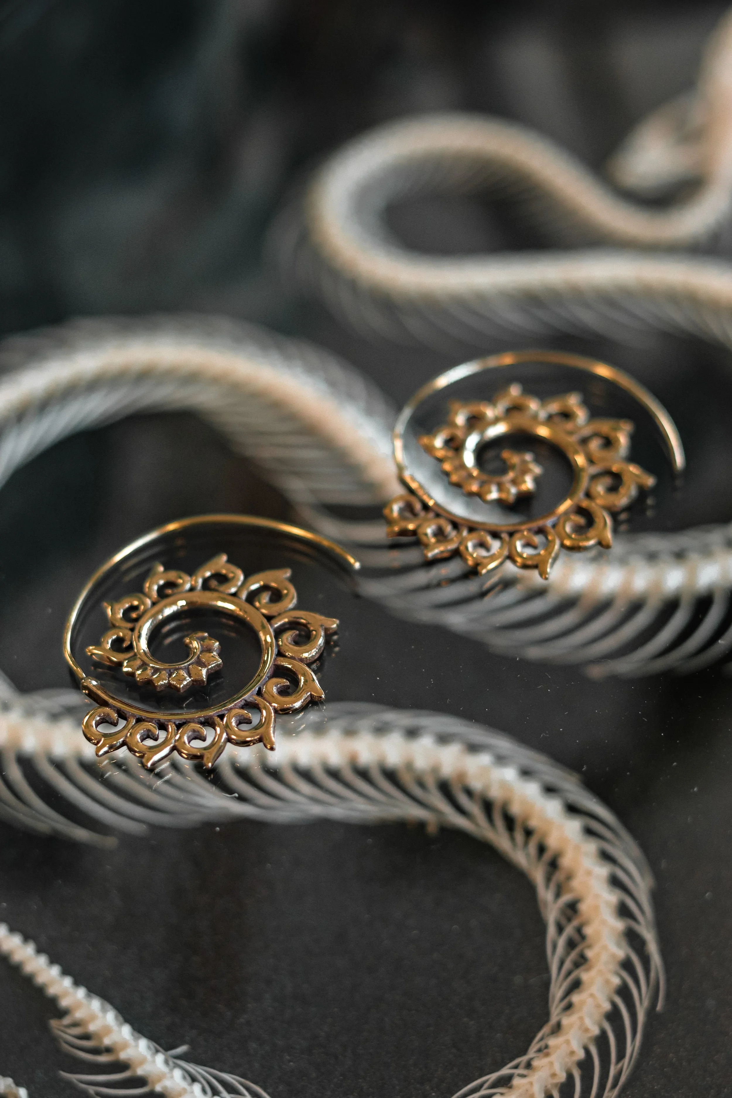 Goudkleurige oorbellen met een spirale en bloemachtig ontwerp, op een zwarte ondergrond met witte gevlochten snoeren.