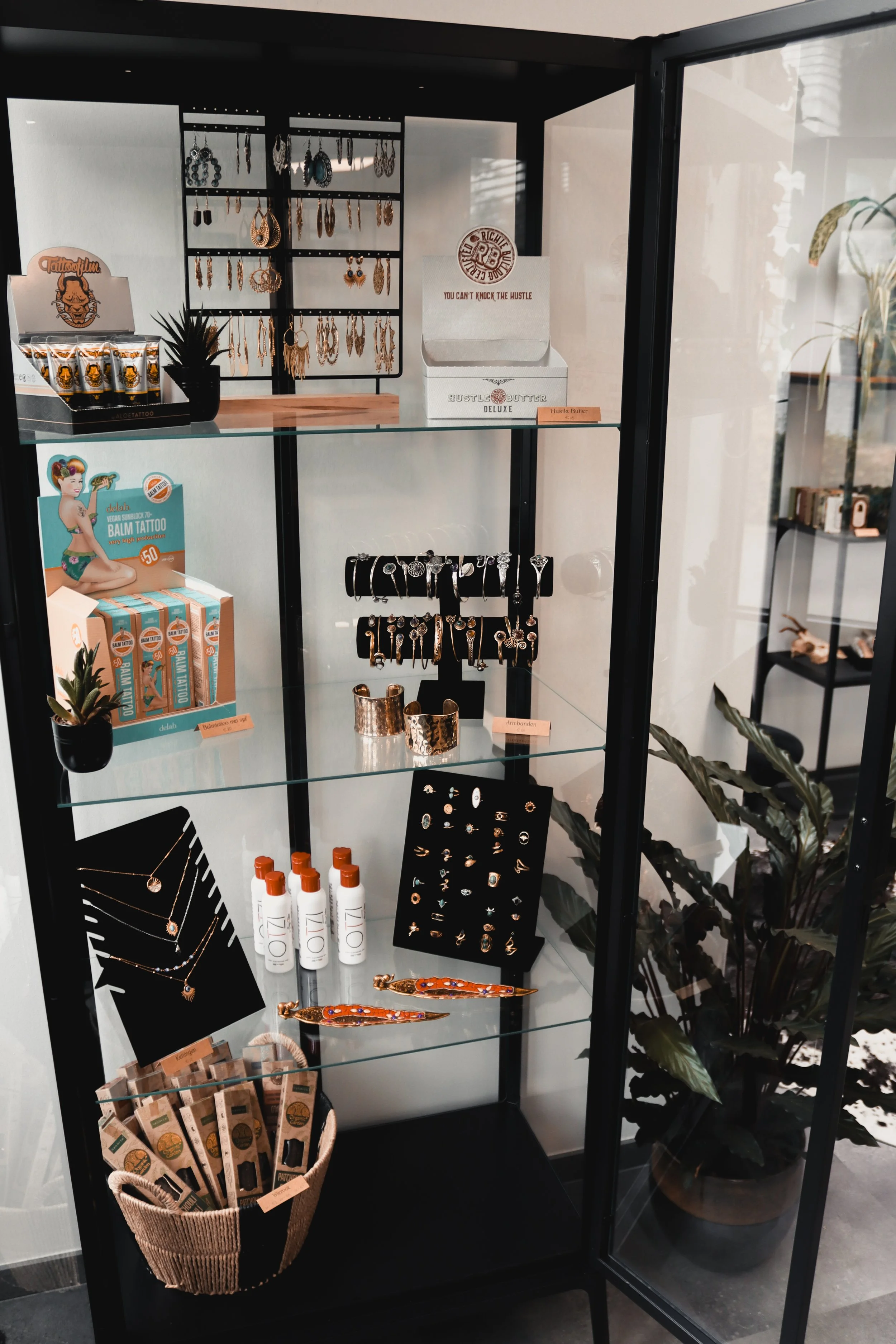 Glasschuifkast met sieraden, huidverzorgingsproducten, en accessoires, met planten op de achtergrond.