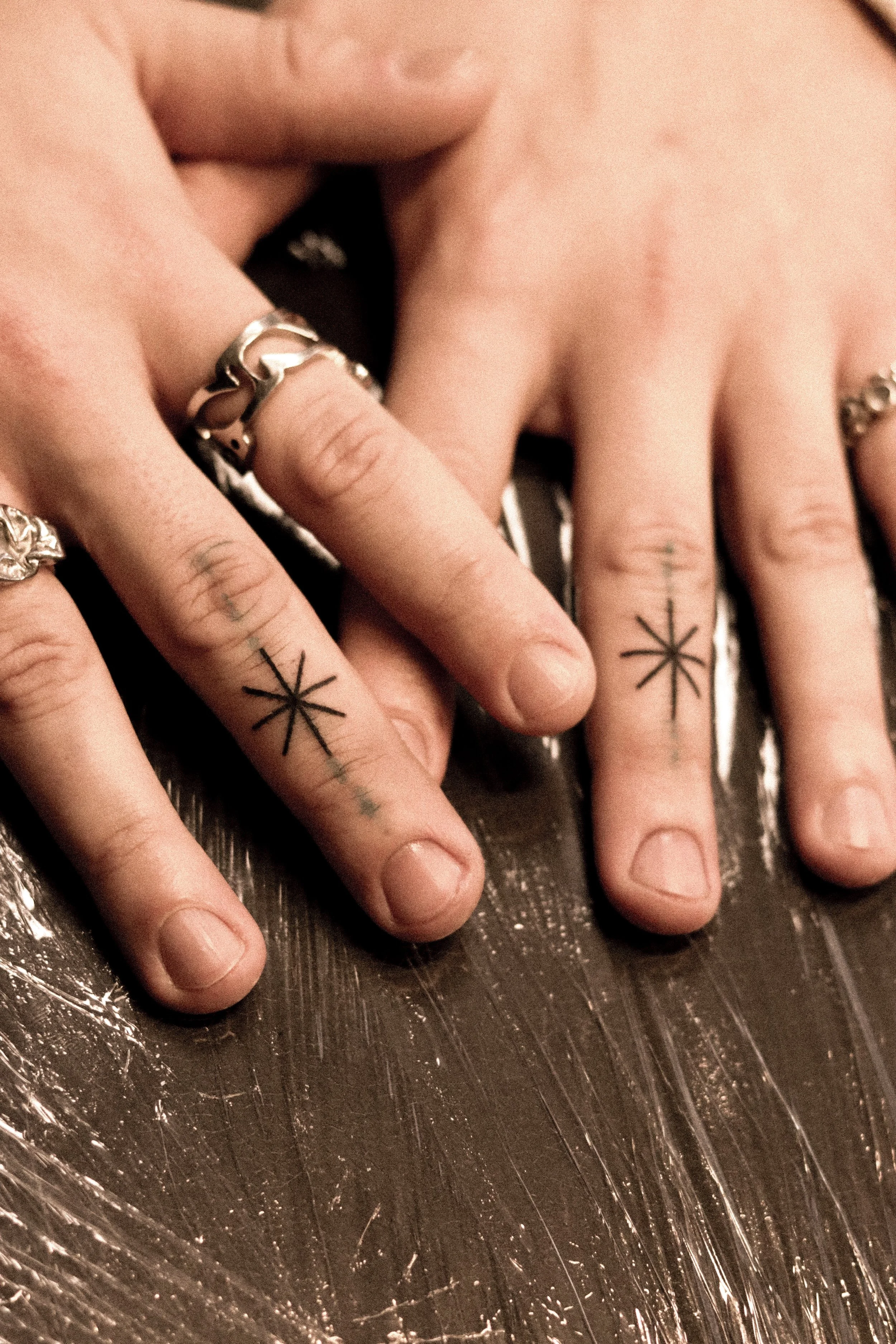 Twee handen met zwarte tatoeages van sterren op de vingers, meerdere zilveren sieraden, rustend op een oppervlak met plastic folie eronder.