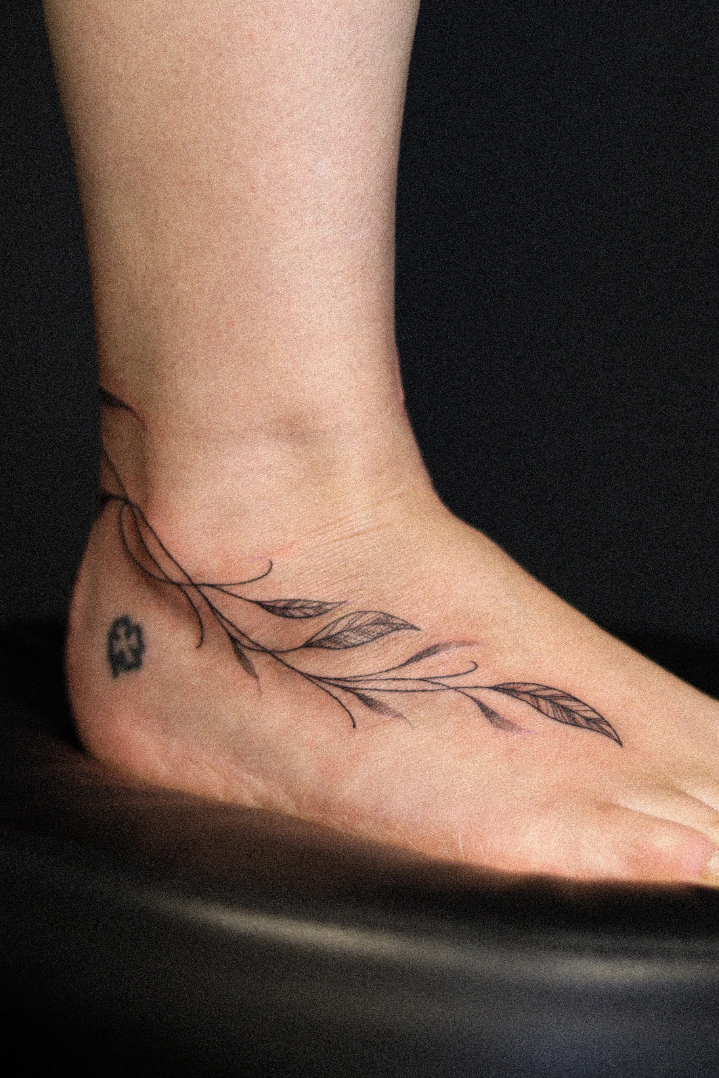 Een voet met een subtiele, lijntekening tattoo van een tak met bladeren en een kleine bloem.