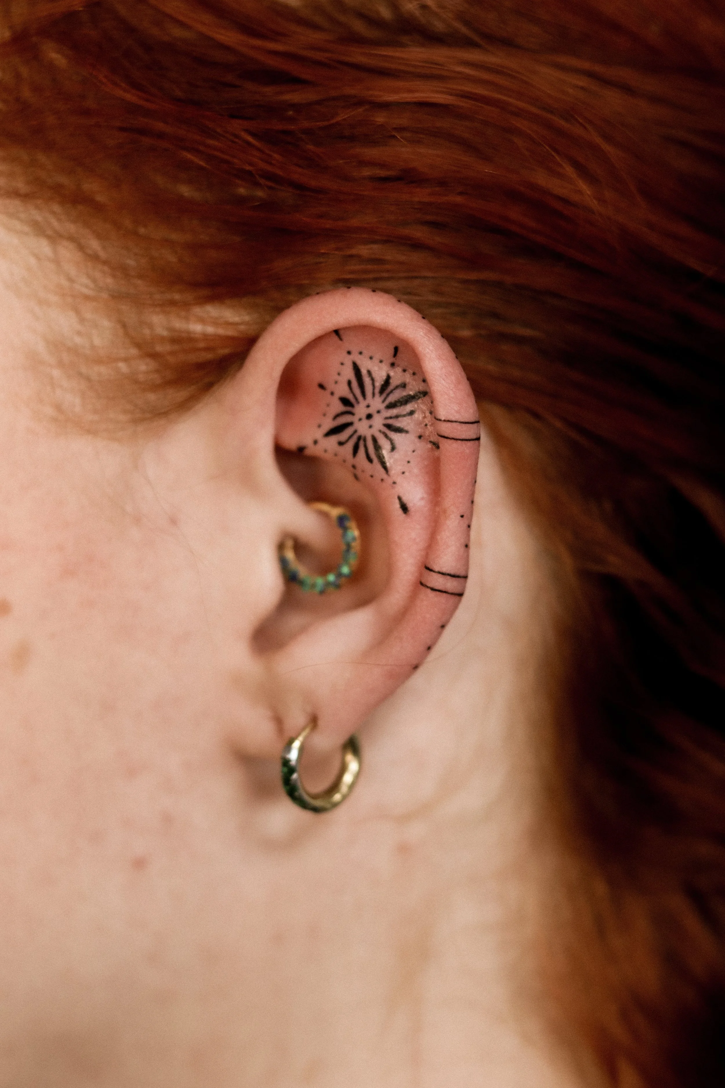 Close-up van een oor met tijdelijke tatoeage van een bloem in het oorkanaal, meerdere oorringen, en zwarte markeringen op de rand van de oor. 