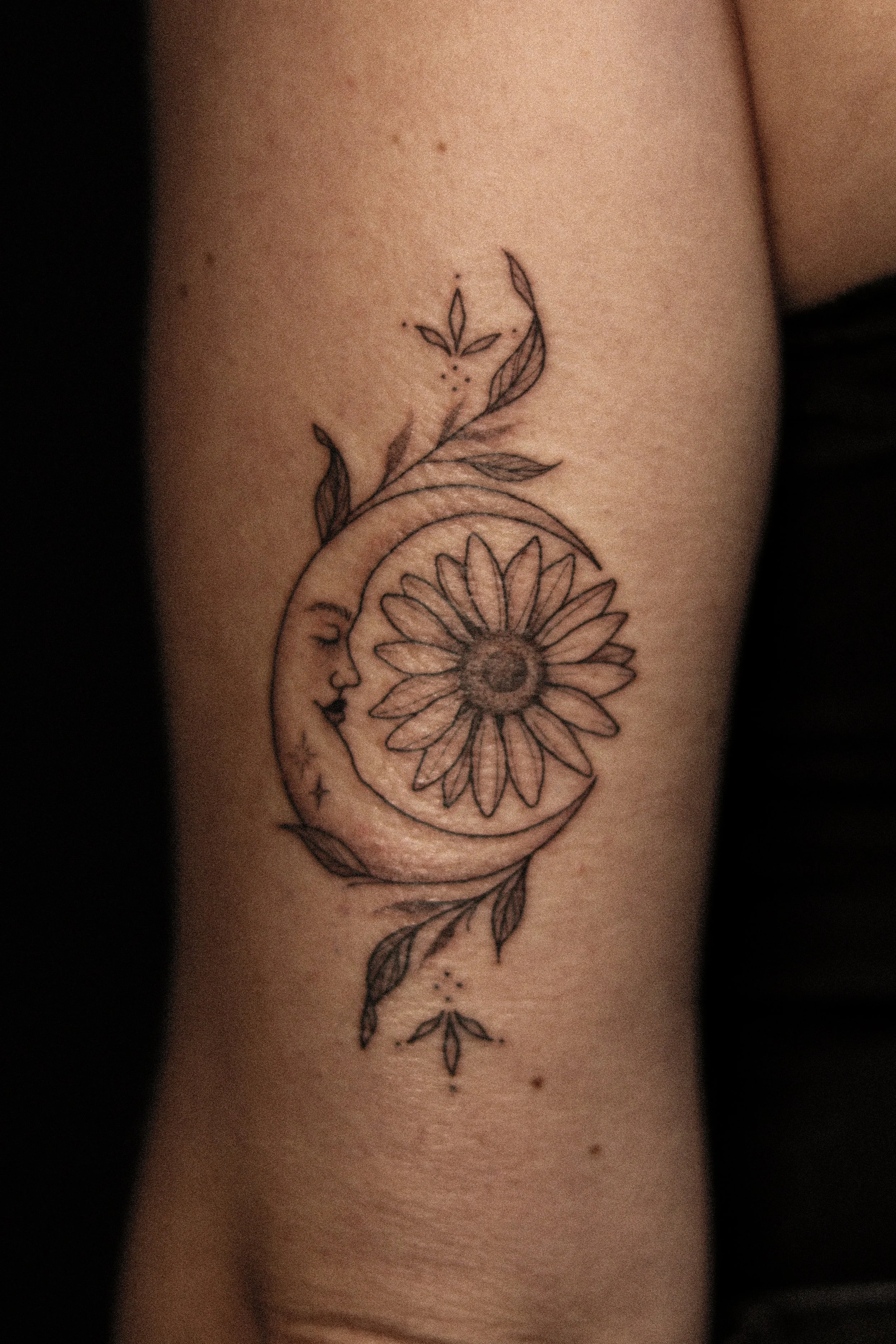 Tatoeage van een lachende maan met een grote bloemen in het midden en bladeren eromheen.
