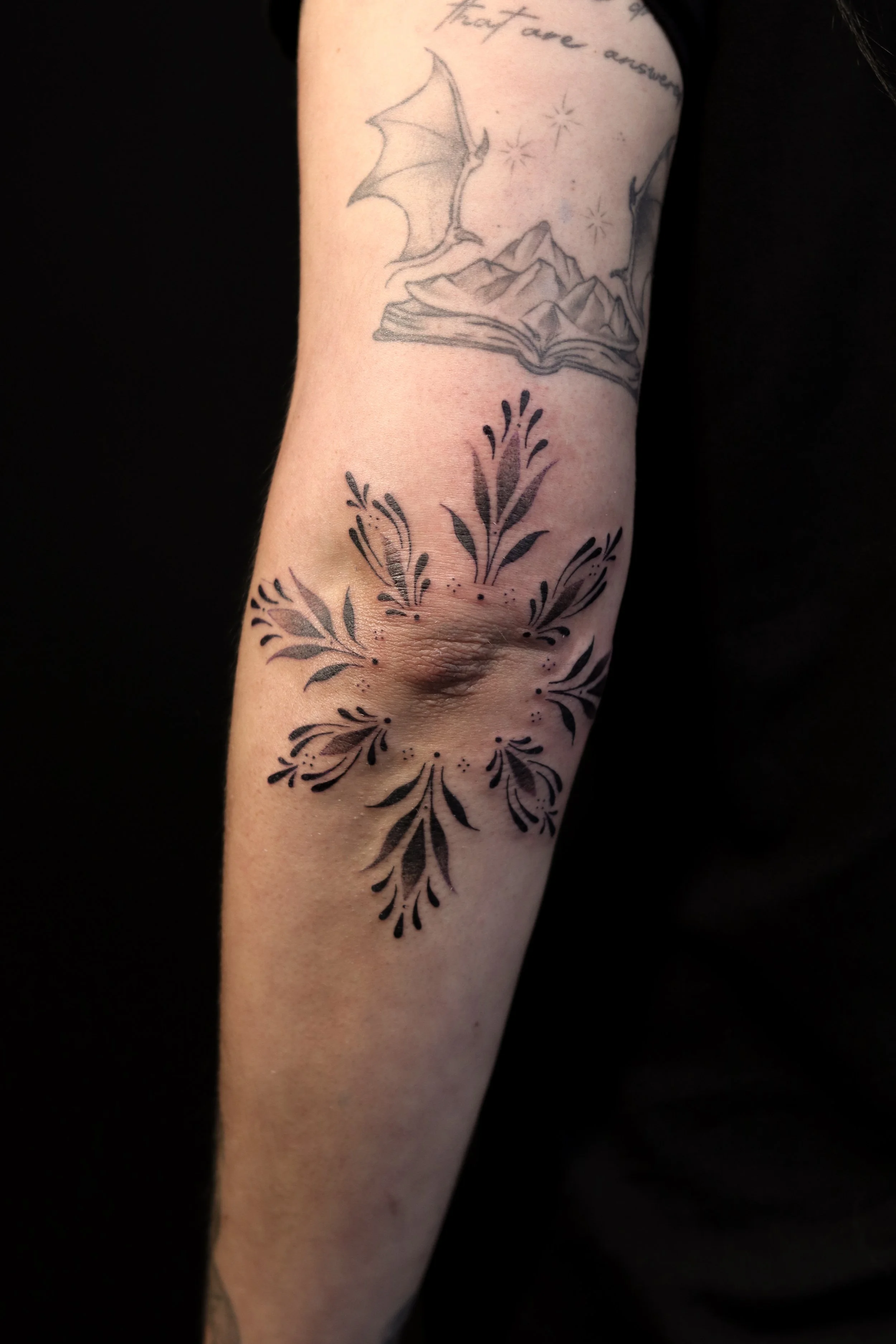 Tatoeage van een vaas met bloemen en een andere tatoeage van een open boek met bergen en vleermuizen er boven, op een arm.