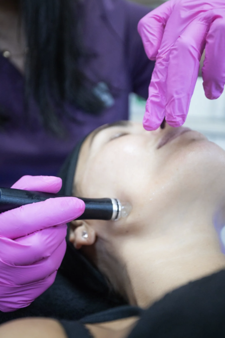 A person receiving a facial treatment or skin care procedure with a professional using a handheld device on their face, wearing pink gloves.