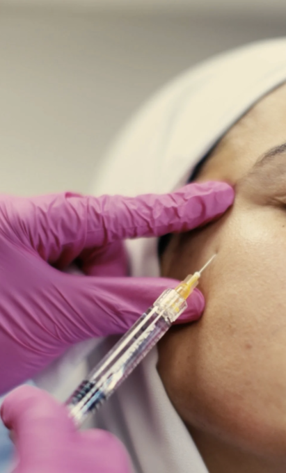 A medical professional wearing pink gloves administering a dermal filler or Botox injection to a patient's cheek.