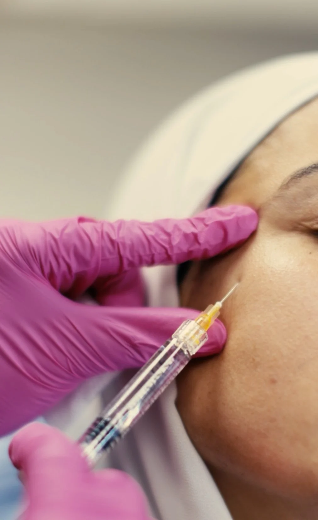 A healthcare professional wearing pink gloves administering an injection into a patient's face, near the eye area.