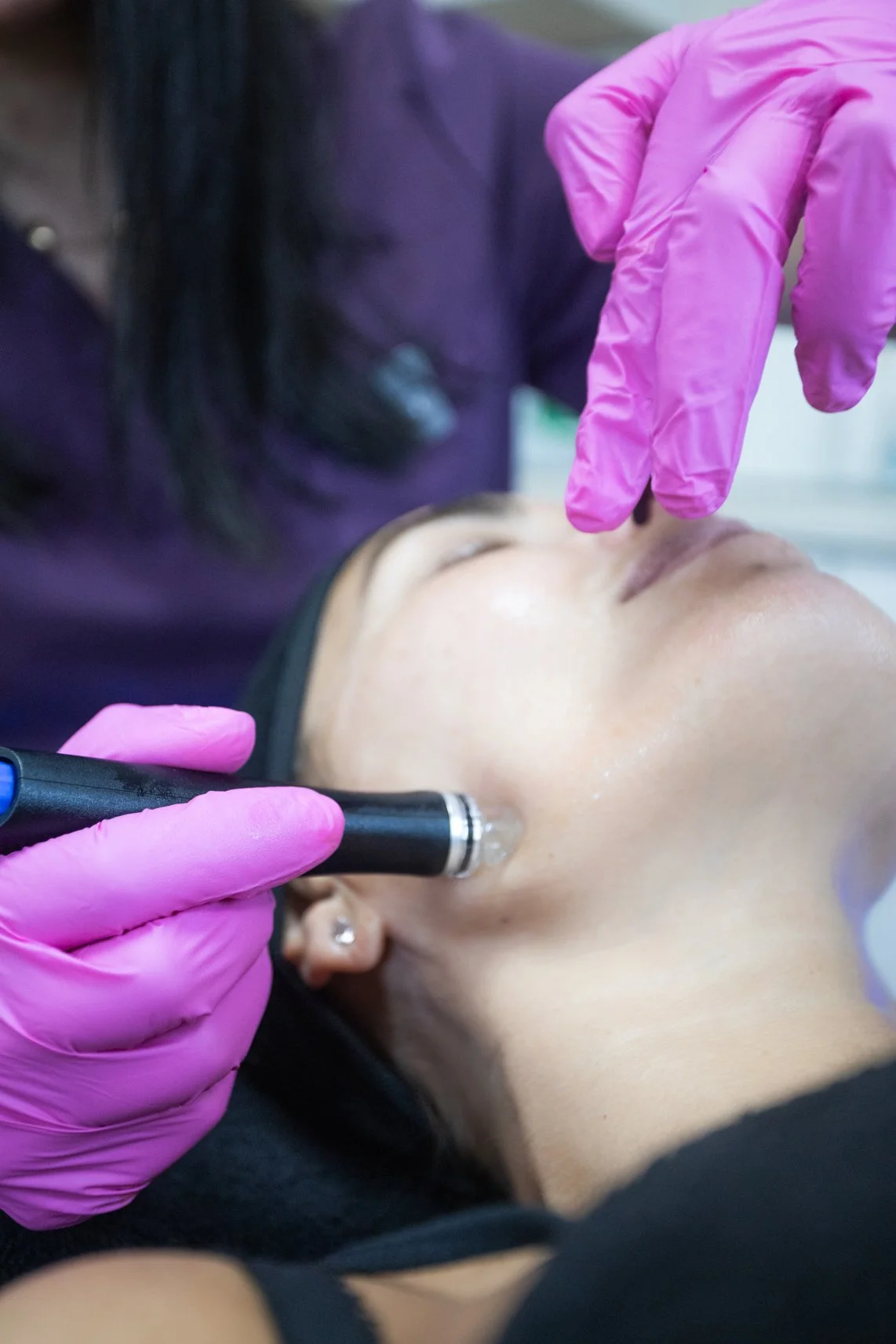 A person receiving a facial treatment with a black device, while a technician wearing pink gloves uses her finger to examine the person's nose.