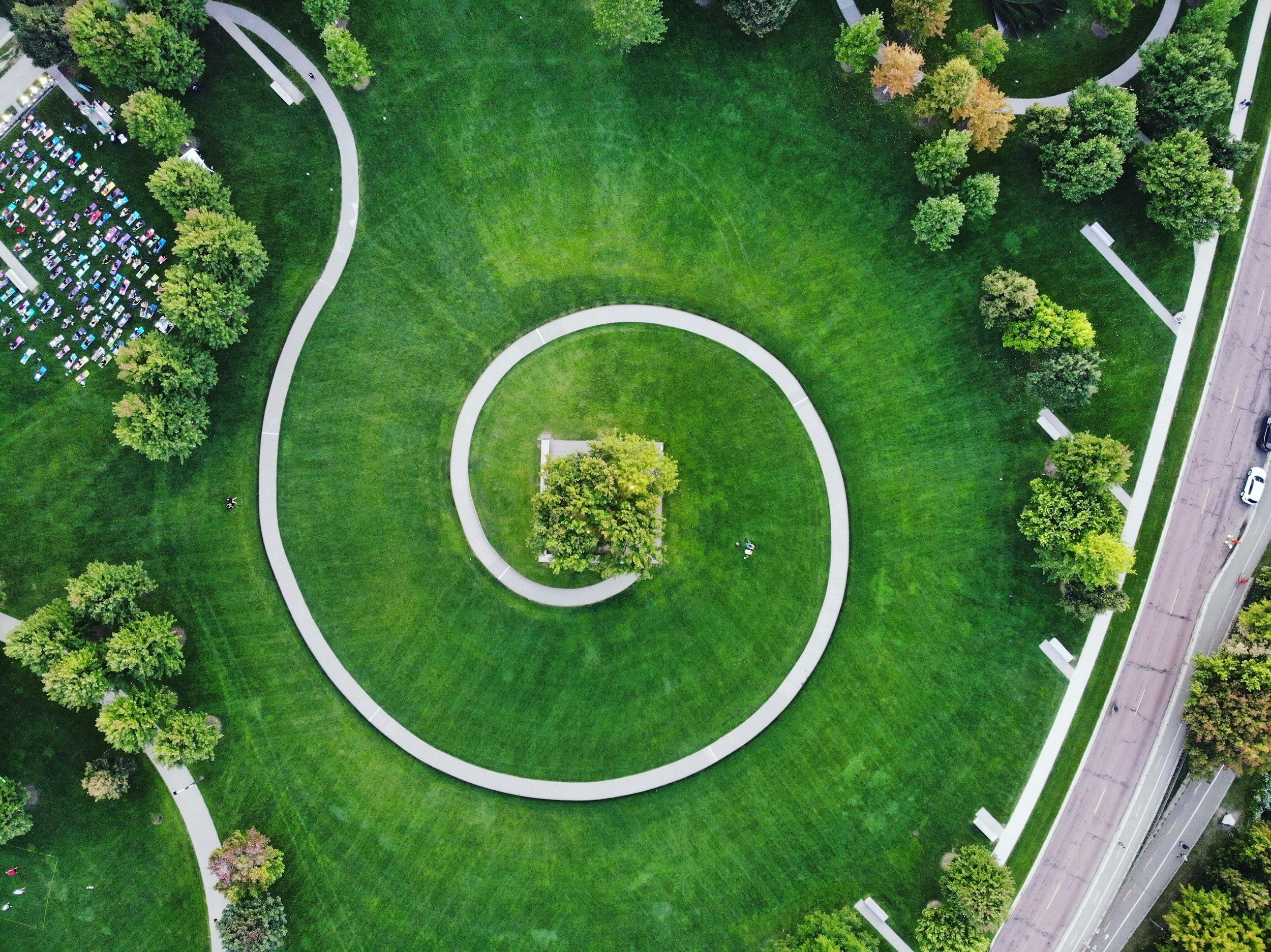 An aerial view of a park with green grass, a winding path, and trees. There is a small building or monument at the center of the spiral path. Parking lot and street are visible at the edges.