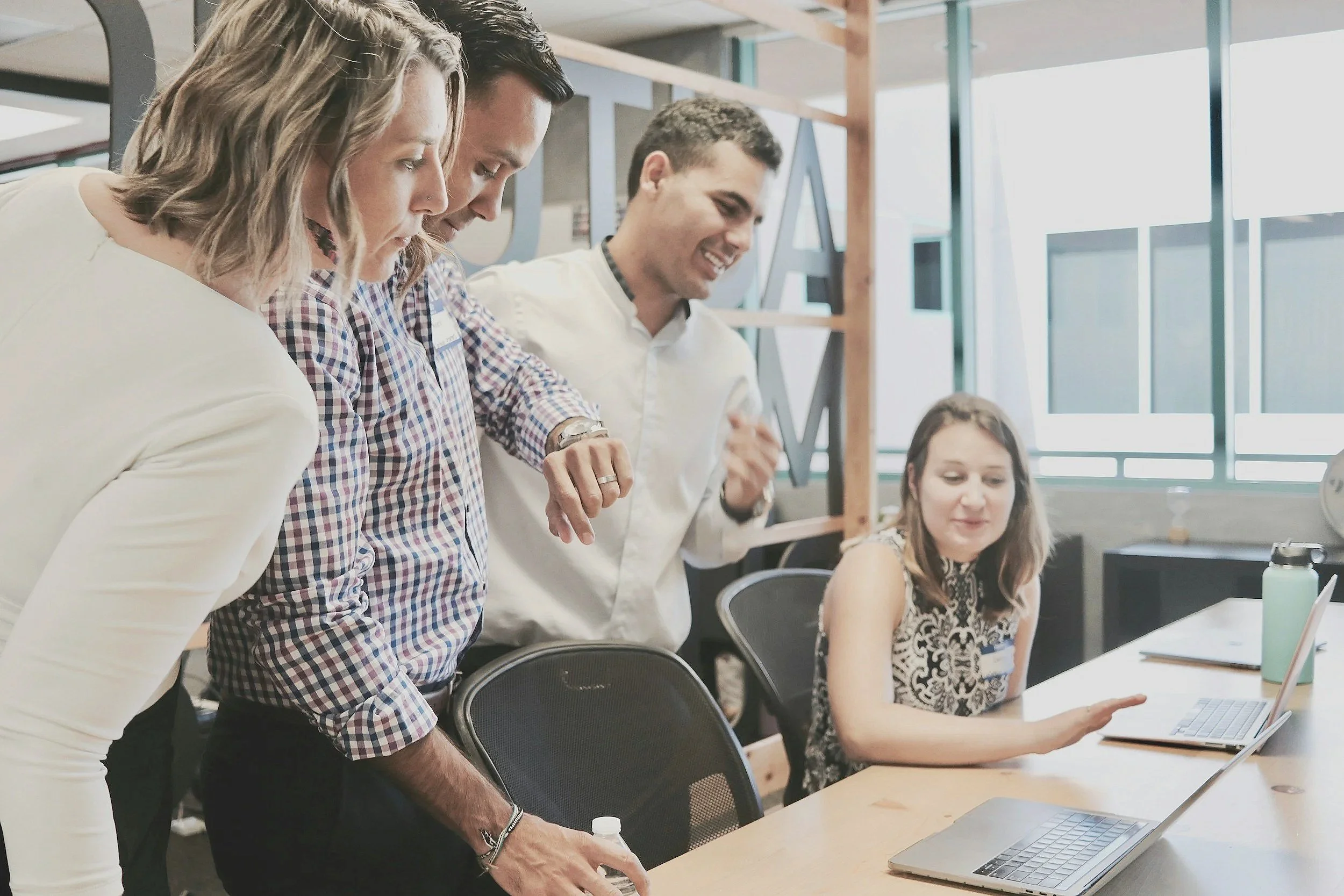 Neljä ihmistä työskentelee yhdessä konttorissa, katsovat tietokoneen näyttöä ja keskustelevat, yksi nainen istuu ja muut seisovat.
