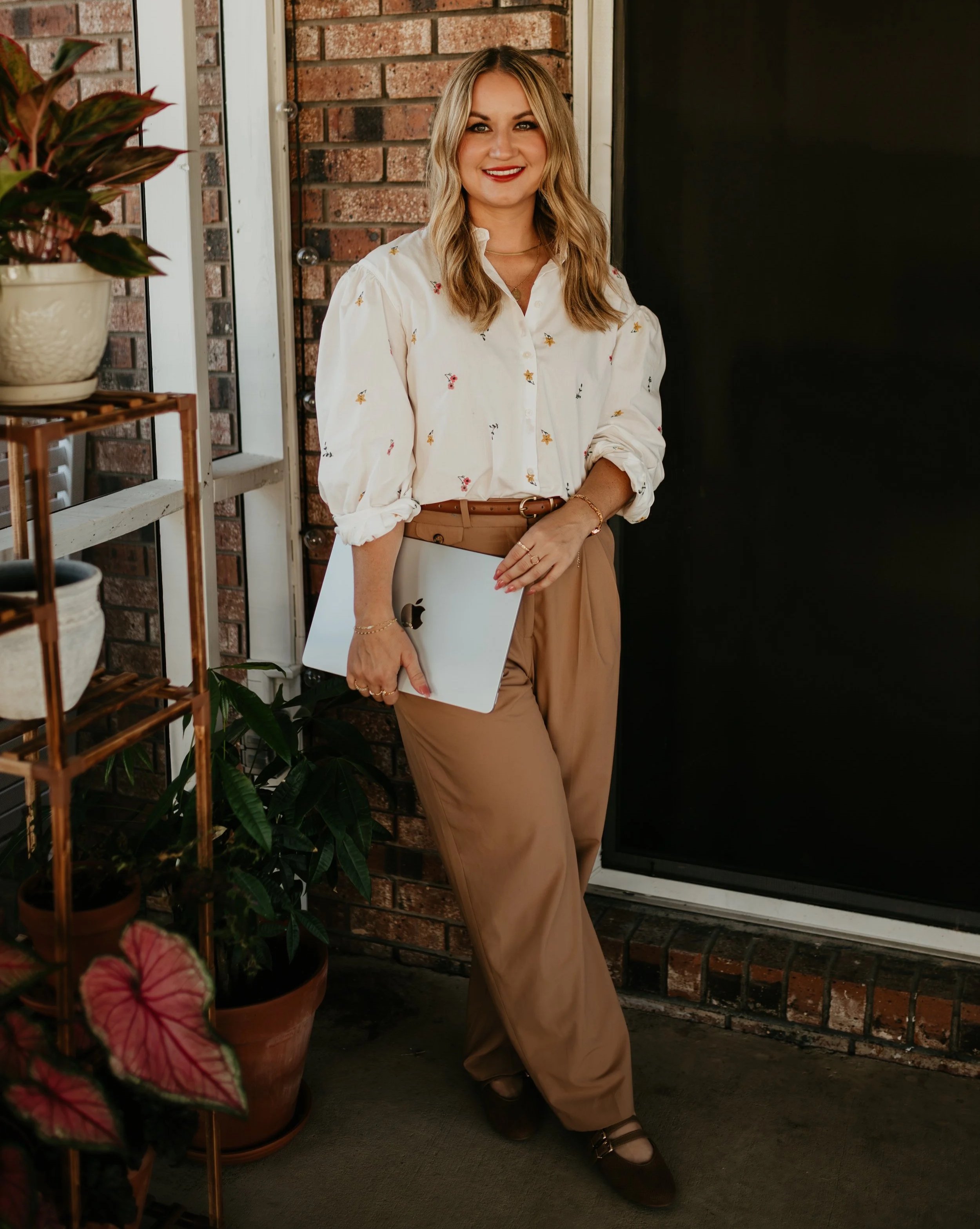 A woman with blonde hair smiling, holding a silver laptop, dressed in a white blouse with small colorful patterns and khaki pants, standing in front of a brick wall and a black door, surrounded by potted plants.