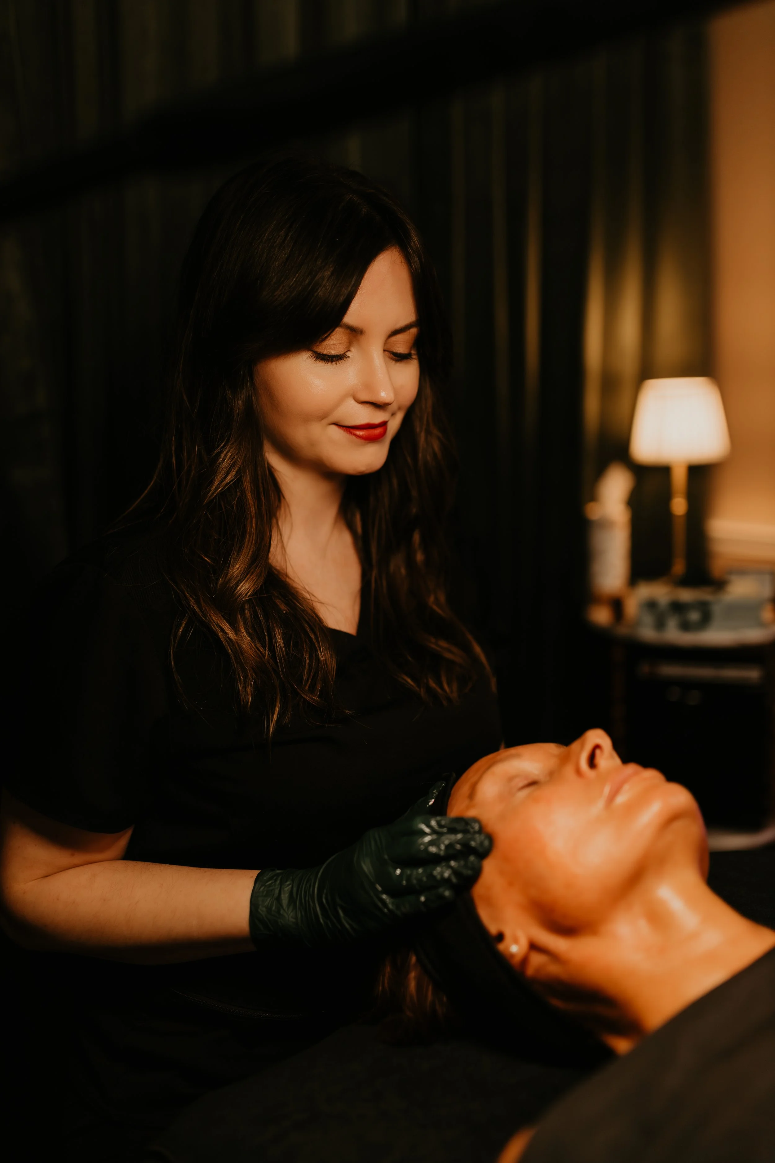 A woman with long dark hair and red lipstick is performing a facial massage or treatment on a woman with red hair who is lying down with her eyes closed. The setting appears to be a dimly lit spa or beauty salon.