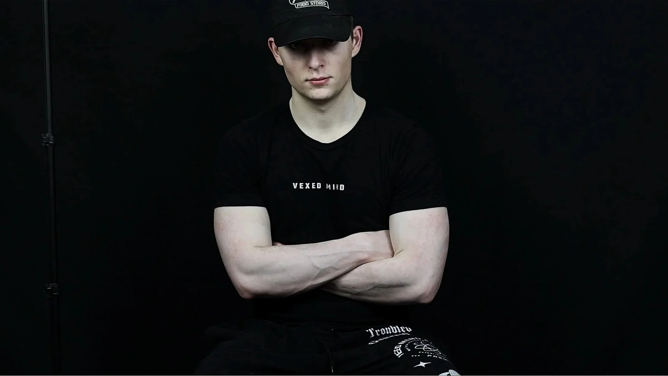 Young man with crossed arms, wearing a black t-shirt with white text and a black baseball cap, sitting against a black background.