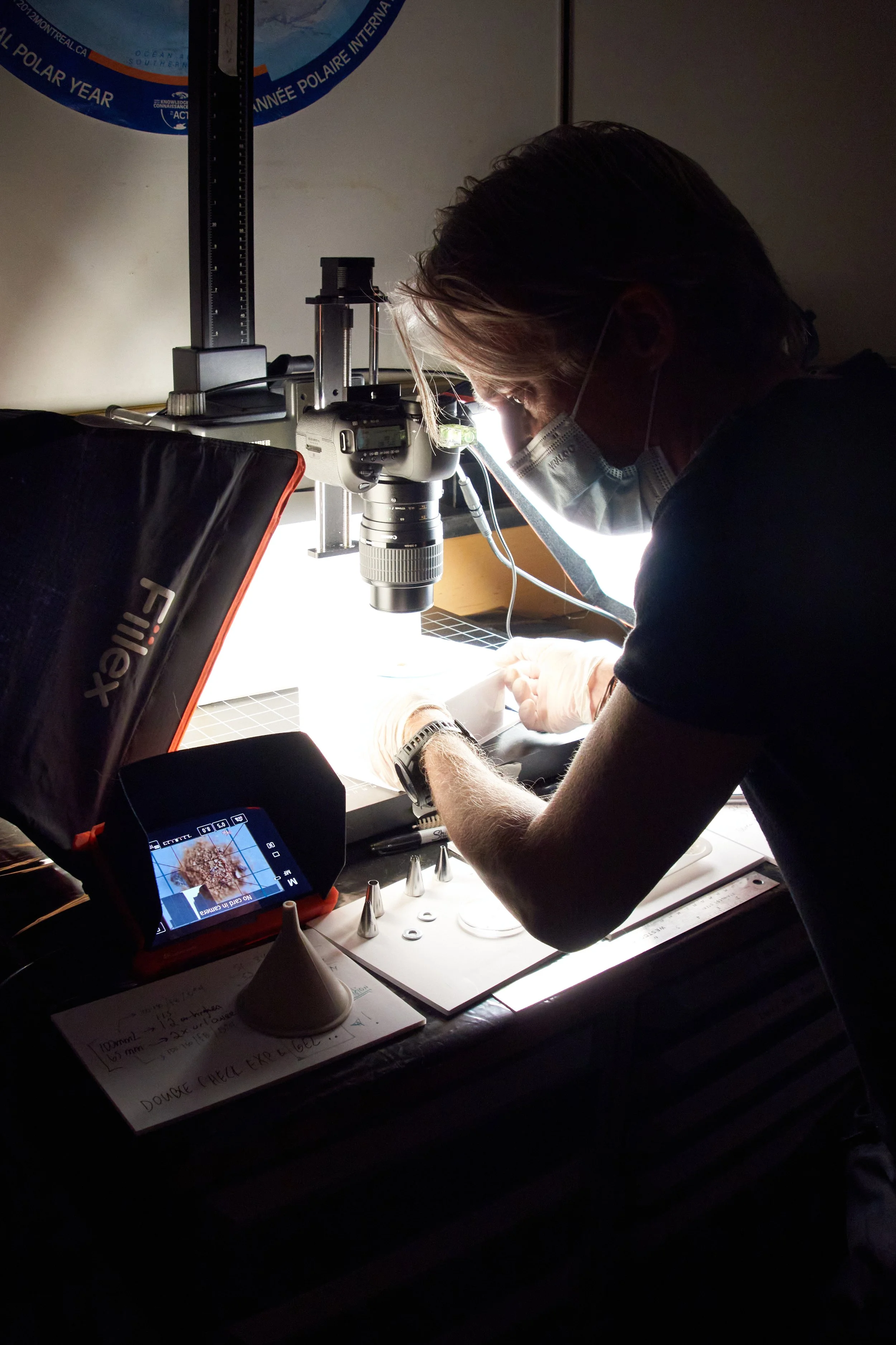 A person wearing a mask and gloves working with a microscope, illuminated by a bright light on a laboratory workbench with scientific equipment and notes.