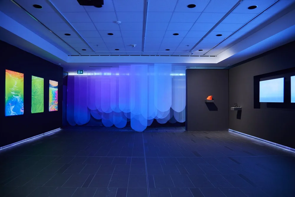 An art gallery room with colorful digital art displays on the left wall, blue lighting projection on the back wall, and a dark tiled floor.