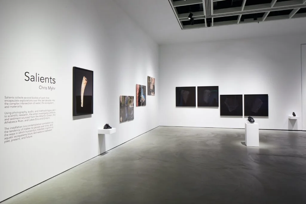 An art gallery showing a series of photographs and artworks on white walls, with small sculptures displayed on white pedestals. The wall on the left has the title 'Salients' by Chris Myhr along with an explanation of the exhibit.