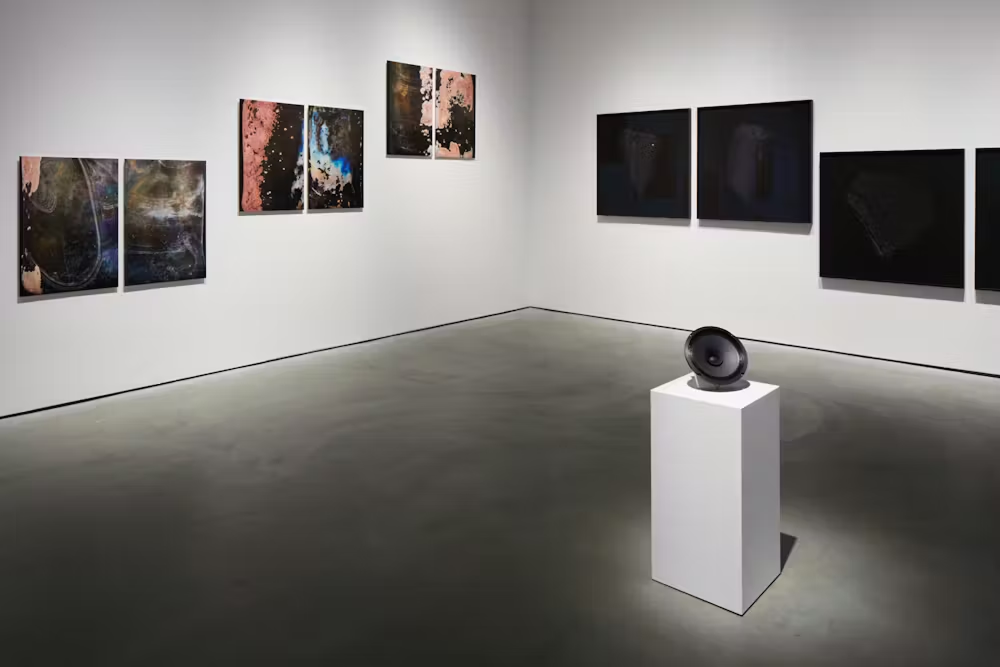 Art gallery with abstract paintings on white walls and a black speaker on a white pedestal.