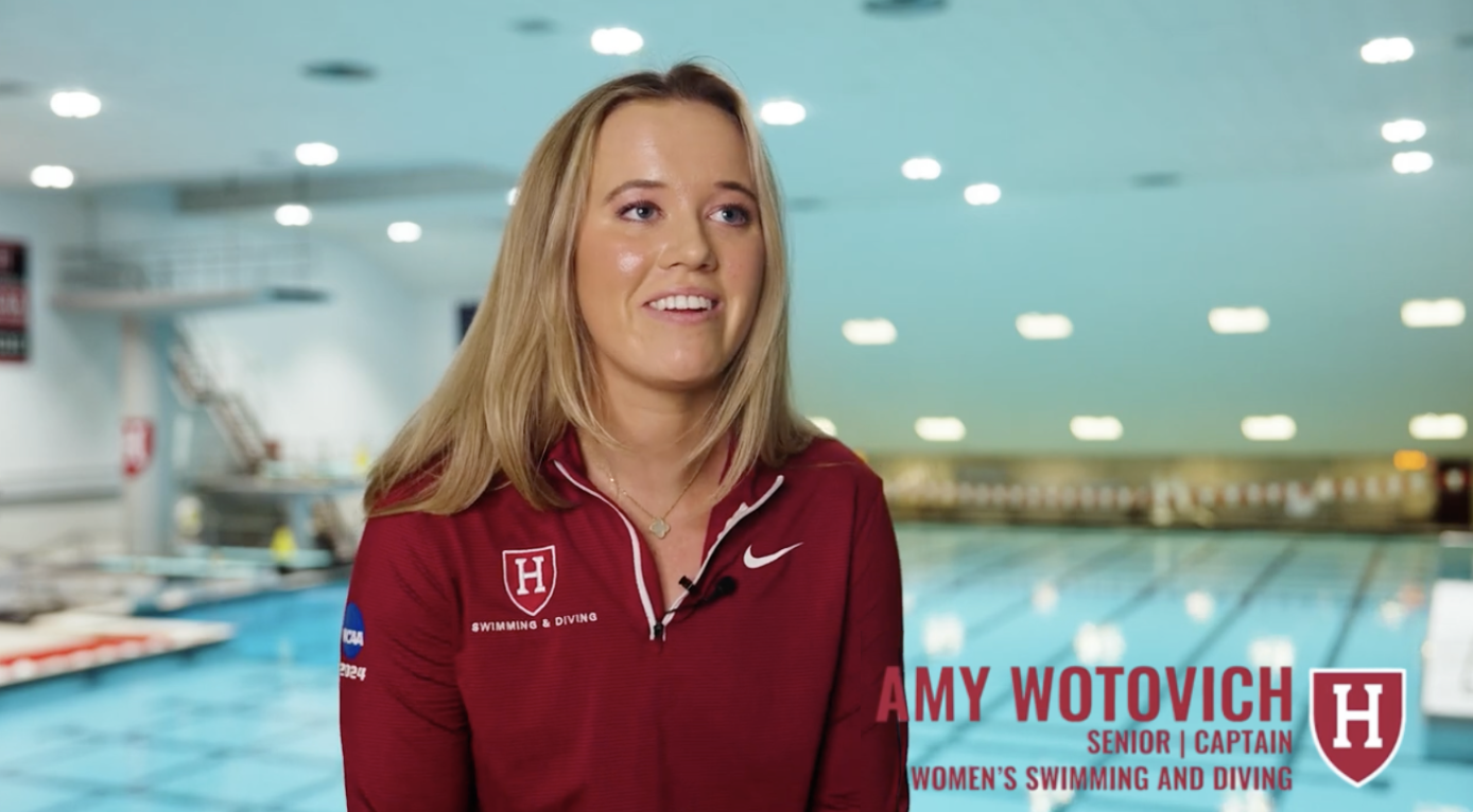 Amy Wotovich, senior captain of the women's swimming and diving team, standing next to a swimming pool in an indoor swimming facility, wearing a maroon Harvard sports shirt with the Harvard logo.