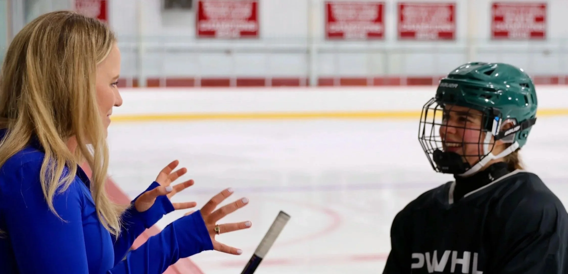 A woman in a blue jacket talks to a young figure skater wearing a green helmet and black figure skating outfit inside an ice rink.