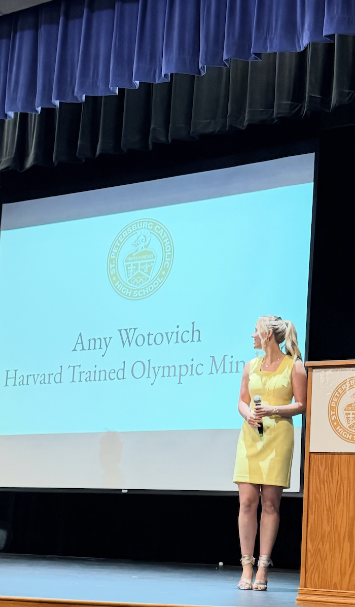 Amy Wotovich standing on stage at St. Petersburg Catholic High School, holding a microphone, with a presentation slide in the background that displays her name and title as a Harvard trained Olympic Mini. The stage has blue curtains overhead, and there is a podium with the school emblem.