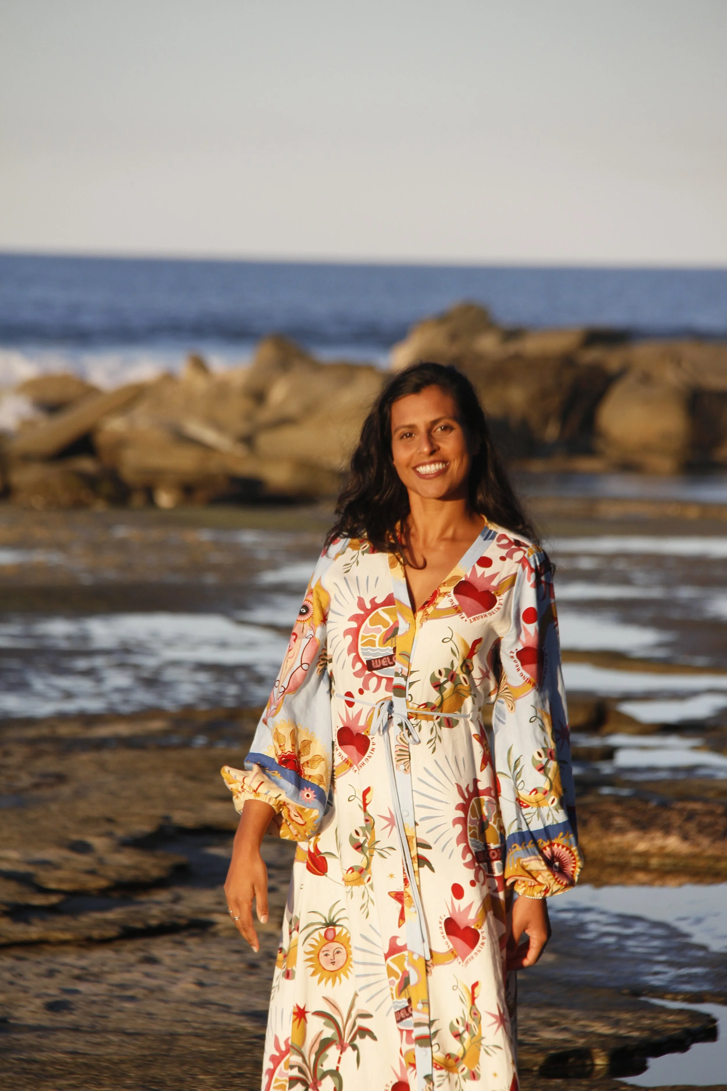 A woman in a vibrant, patterned dress stands barefoot on a rocky shoreline, the ocean stretching behind her under a clear sky. She is still, strong, and fully present — a moment of feminine grounding and connection to the elements.