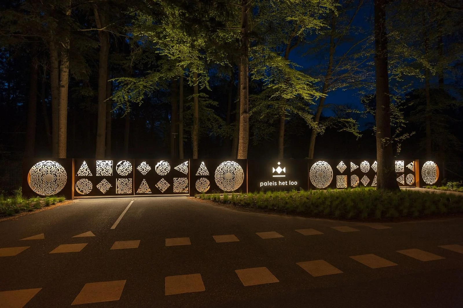 Nighttime view of an illuminated decorative gate with intricate patterns, surrounded by tall trees, with the words 'paleis het loo' displayed on the right side.
