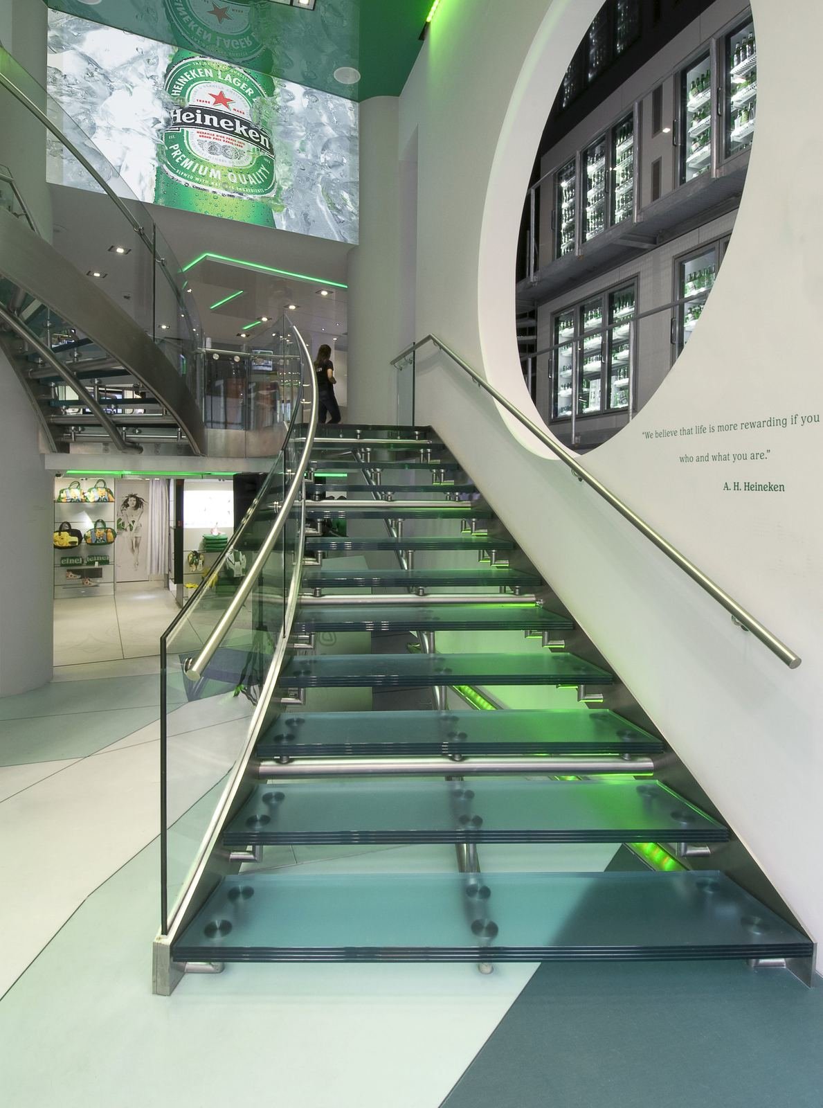 Modern interior with spiral staircase, glass steps, and metal handrails. Visible green wall art and people conversing near large windows.
