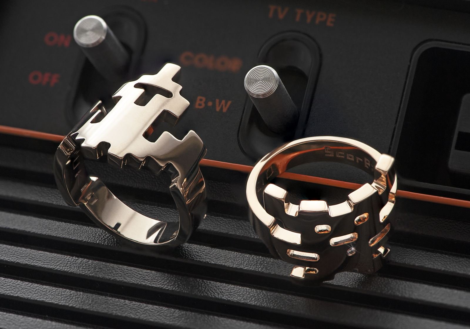 Two silver rings with gear-like designs resting on a black textured surface in front of a control panel with switches and knobs. 	
Oyukio rings Aiko and Kappa on vintage Attari console.