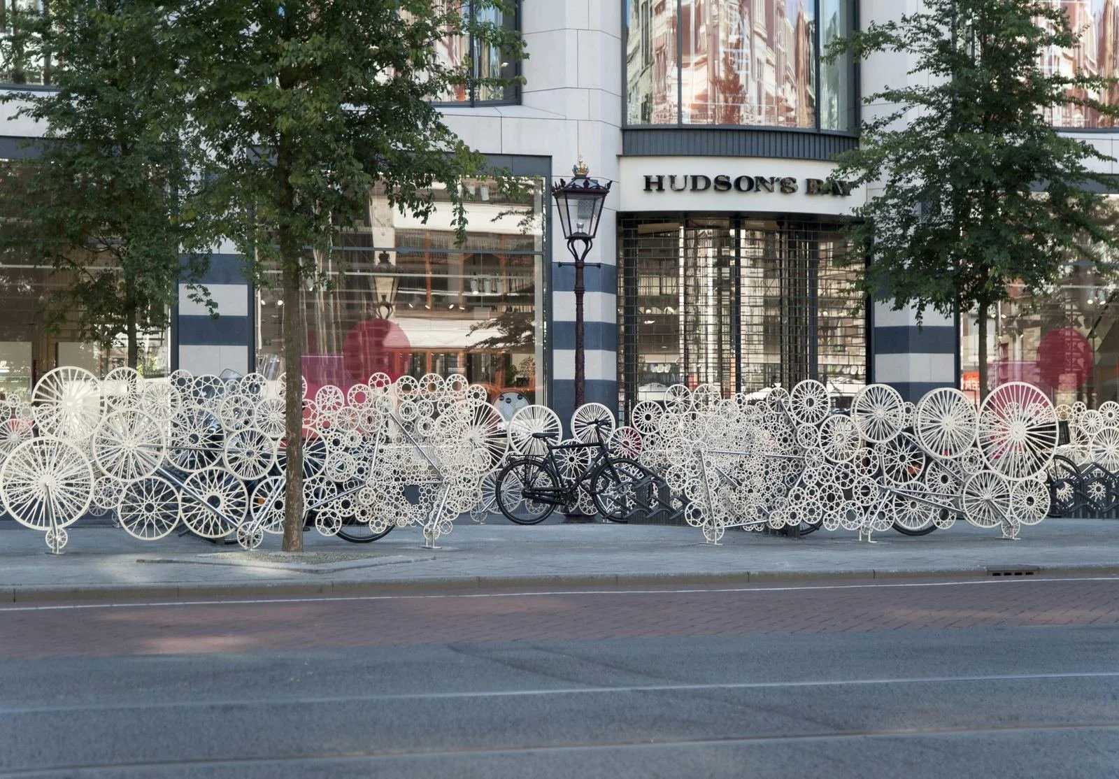 City sidewalk with decorative white bike-shaped sculptures and two trees in front of a building with large glass windows and a sign that reads 'Hudson's Bay'.