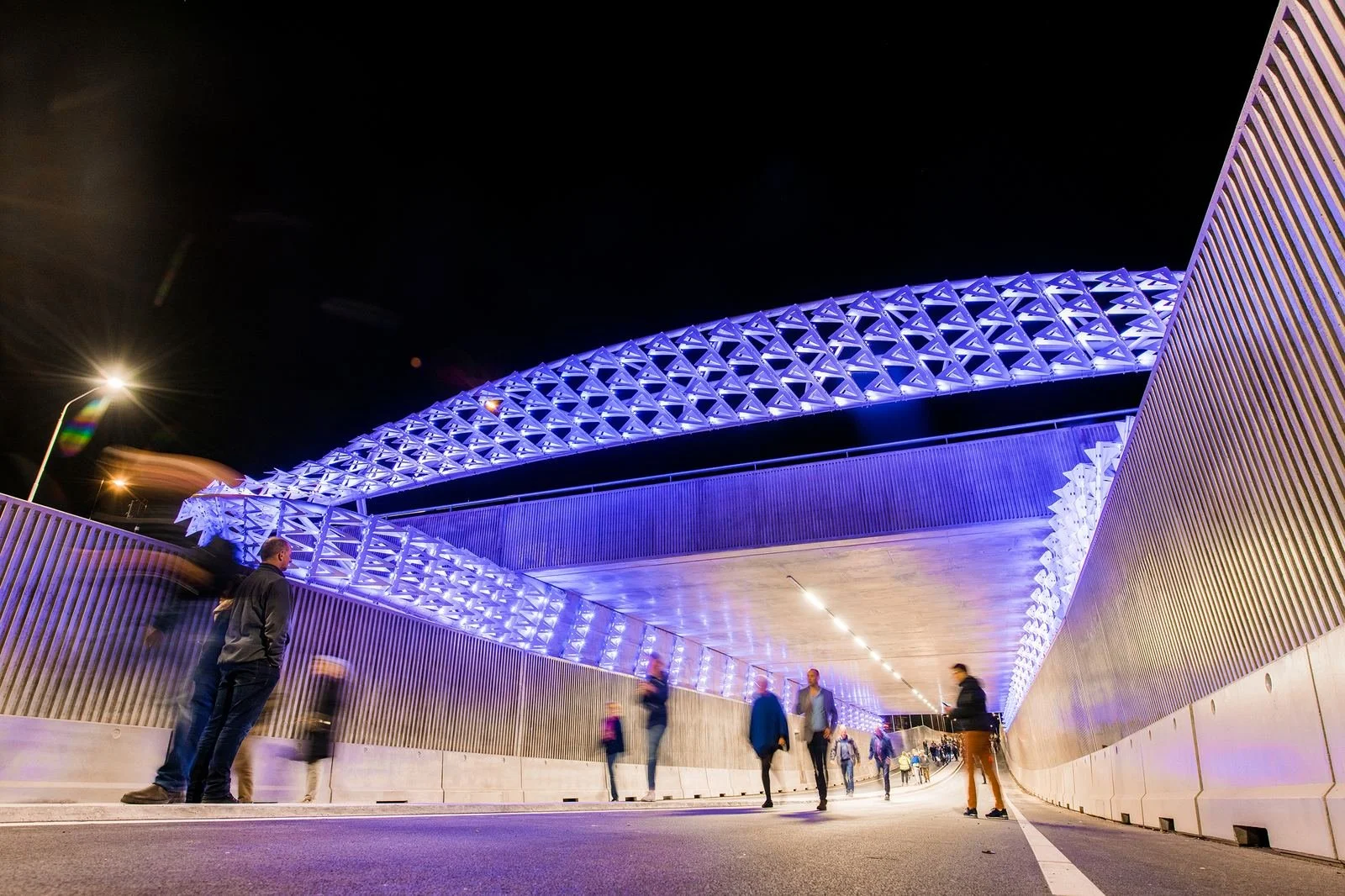 Night view of a modern illuminated pedestrian bridge with purple lighting, metal lattice design, and people walking underneath.