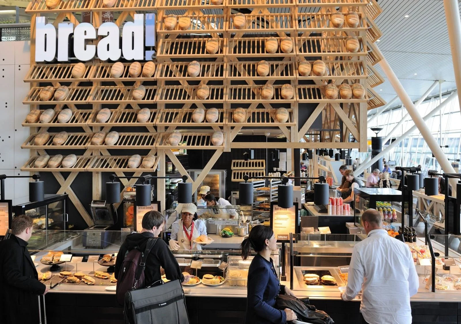 Inside a modern bakery or café, with a large wooden display structure resembling a crate filled with bread rolls, and a sign that says 'bread!'. Customers are ordering food at the counter, and there are various baked goods and beverages on display.