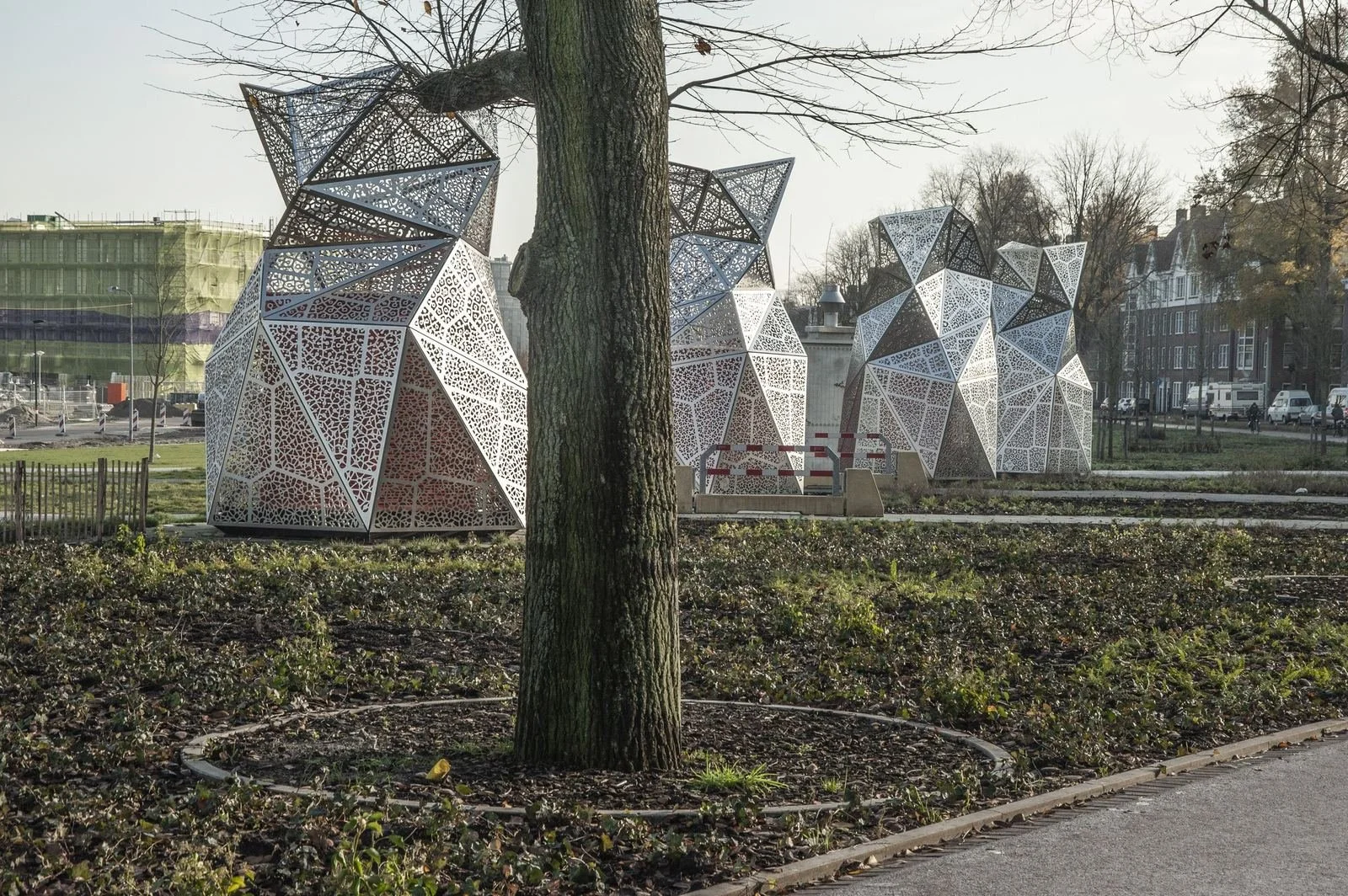 Three modern geometric sculptures made of white perforated metal in a park, with trees, grass, a sidewalk, residential buildings, and a street in the background.