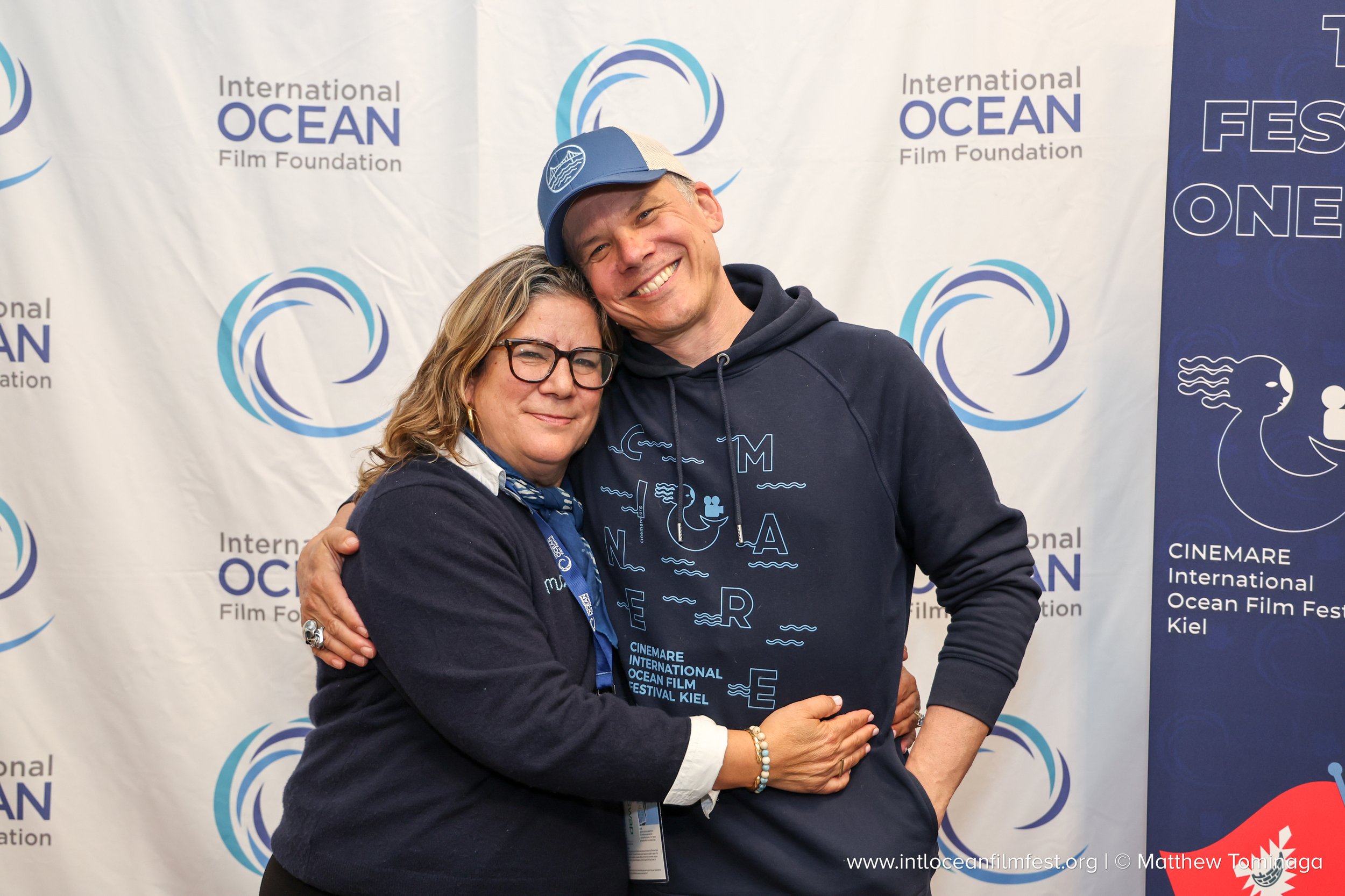 A man and woman hugging in front of a backdrop with logos of the International Ocean Film Festival and other ocean-themed graphics. The man is wearing a blue cap and hoodie, and the woman has glasses and is wearing a dark jacket.