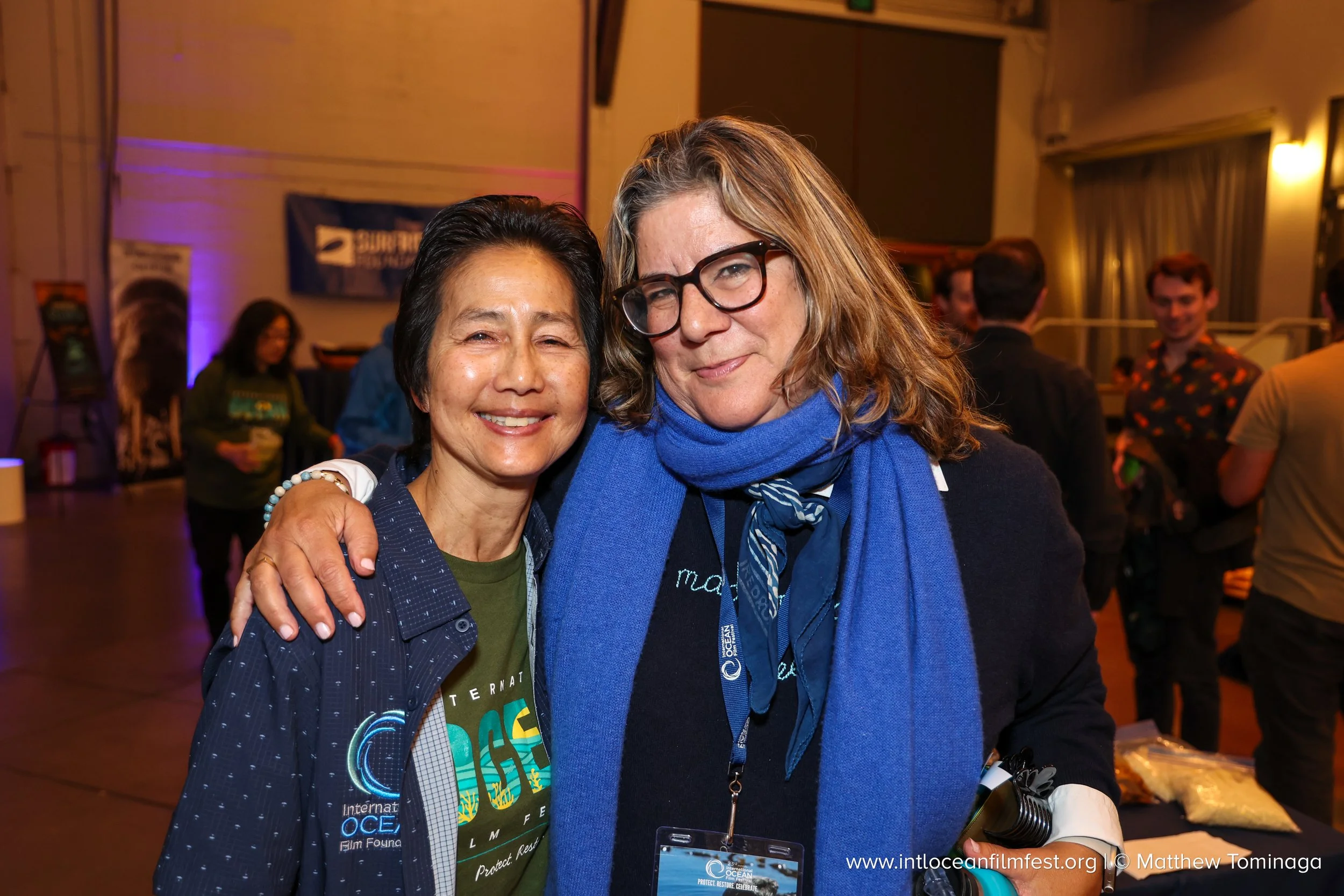 Two women smiling, standing closely with arms around each other at an indoor event, with people in the background. One woman has short dark hair and wears a dark blue jacket, the other has long wavy light hair, glasses, and a blue scarf.