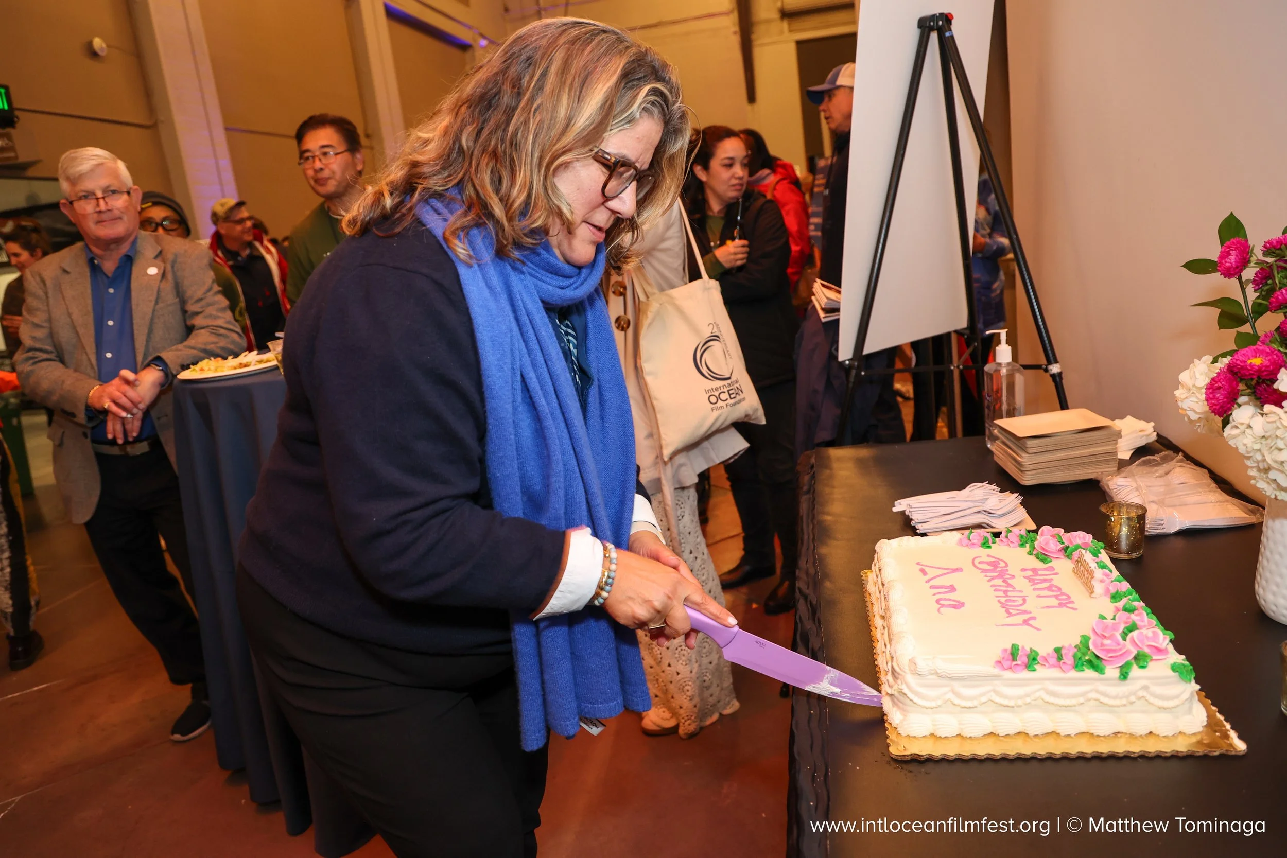 A woman with glasses and a blue scarf is cutting a decorated cake at a celebration or event. Several people are in the background, and there are flowers and a stack of papers or napkins on the table near the cake.