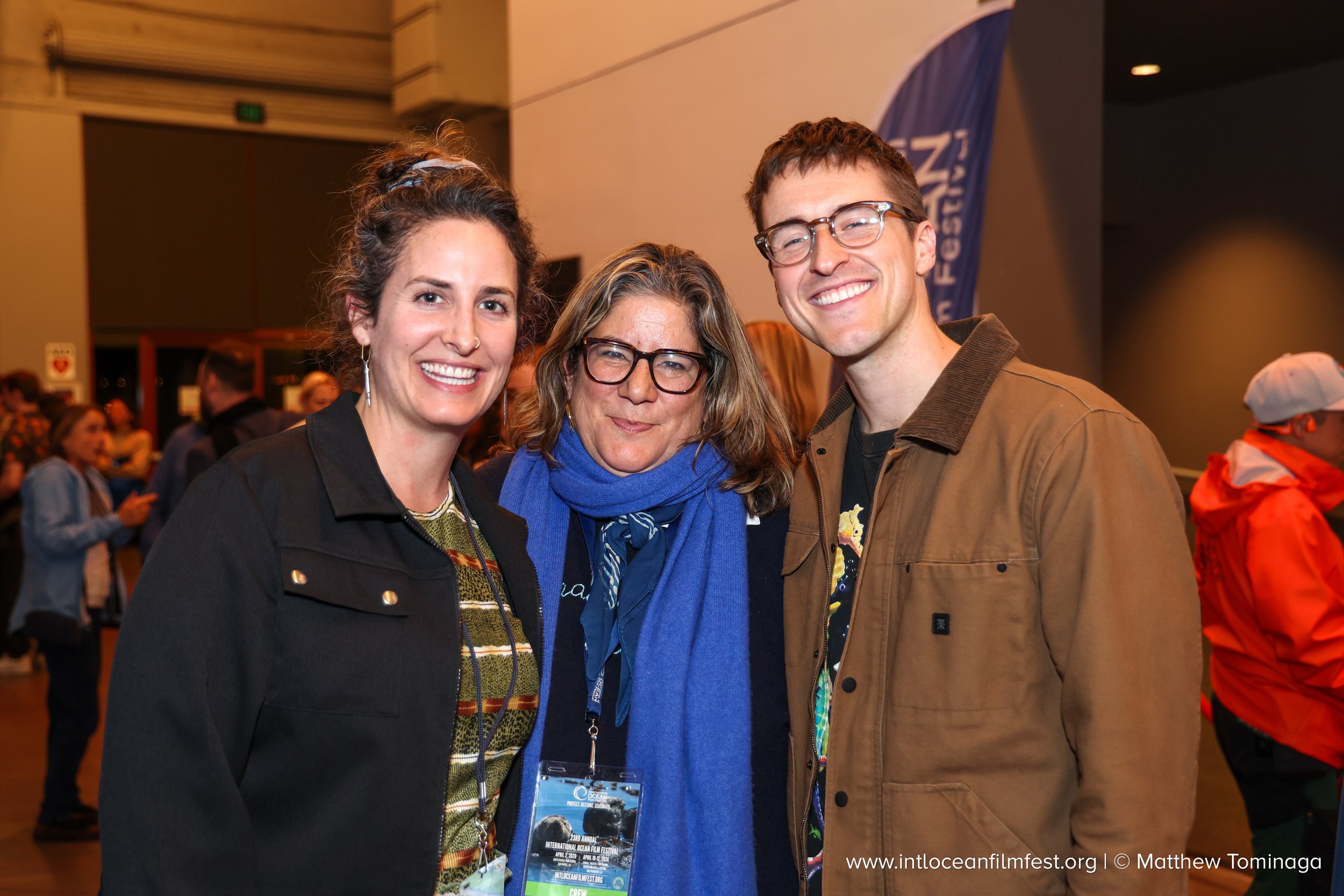 Three smiling people at an indoor event, posing for a photo. The woman on the left has curly hair and is wearing a black jacket. The woman in the middle has glasses and wavy hair, wearing a blue scarf. The man on the right has short hair and glasses,