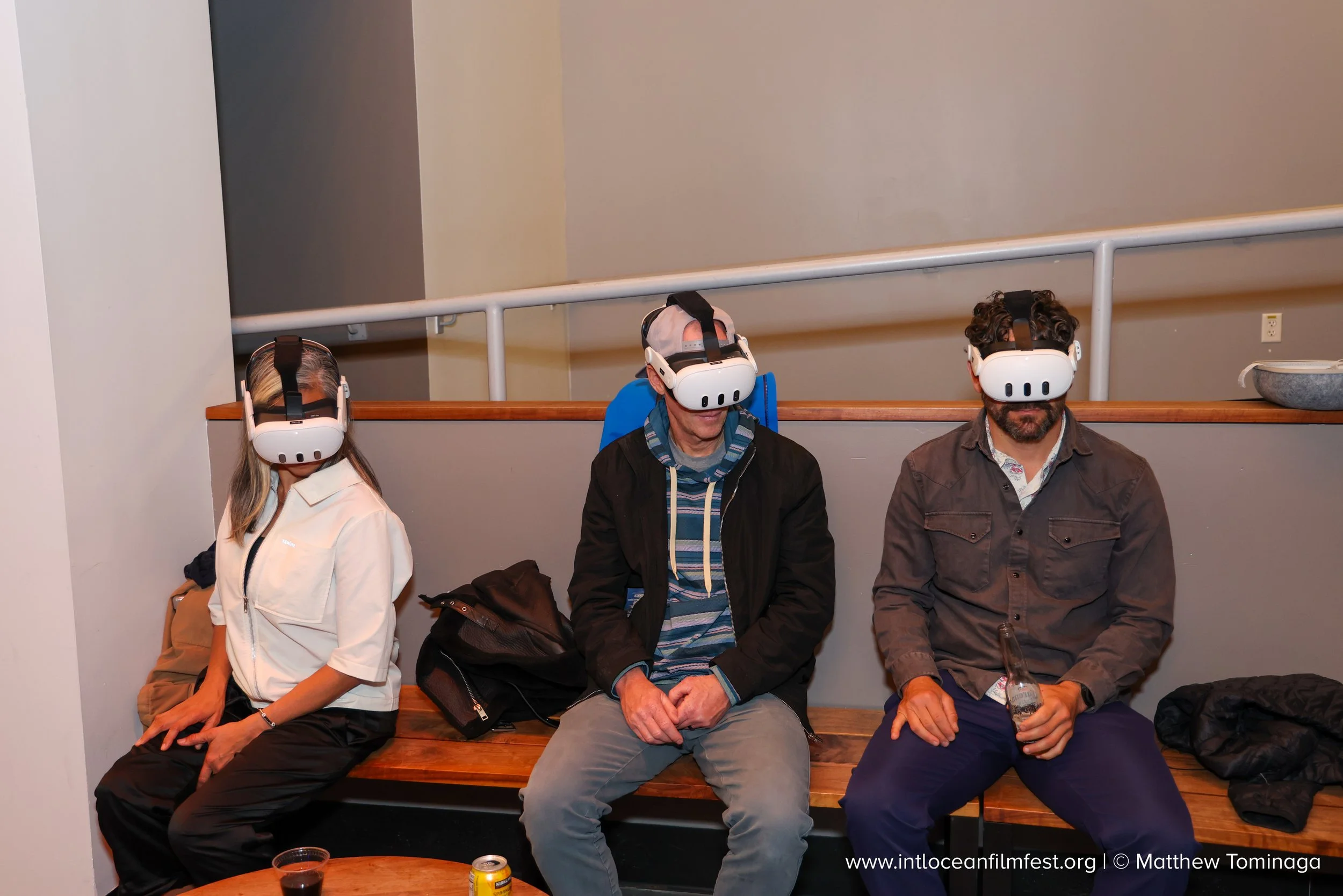Three people sitting on a wooden bench, all wearing virtual reality headsets. The woman on the left has long hair, the man in the center has gray hair, and the man on the right has dark curly hair. Bottles and drinks are on the table in front of them