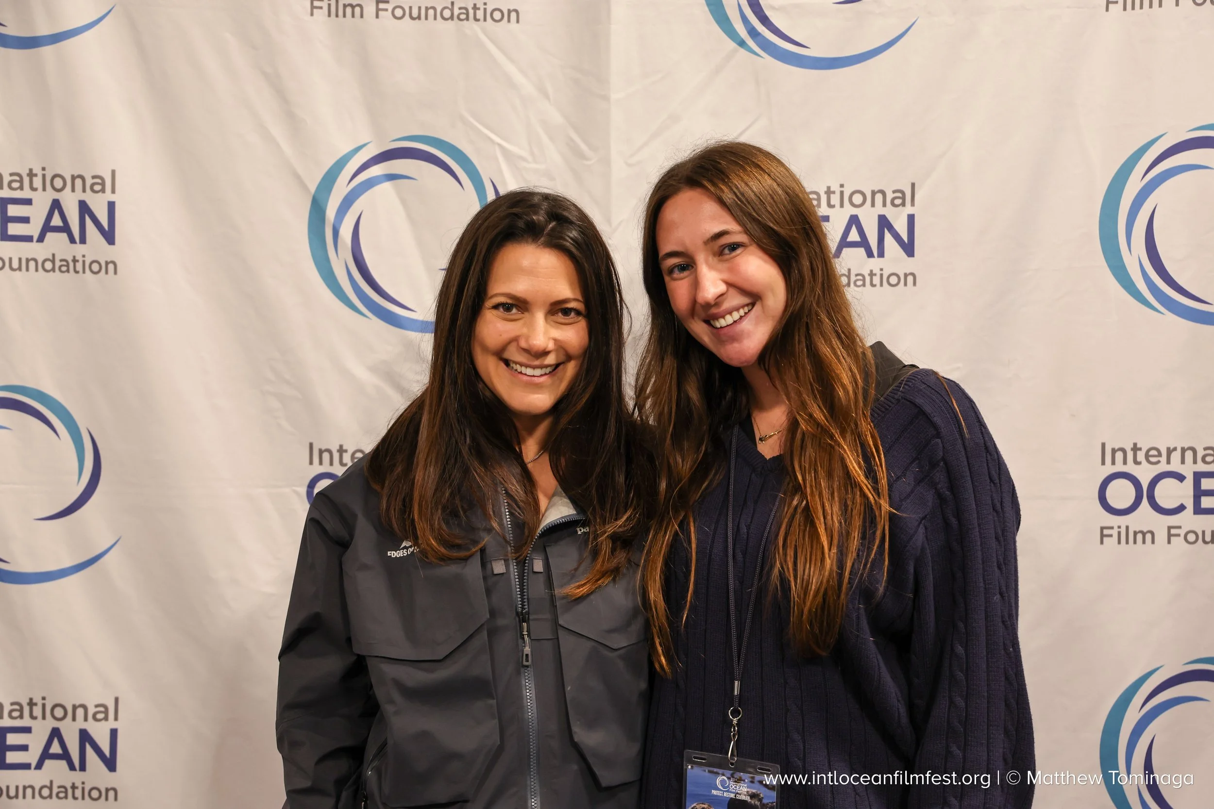 Two women smiling and posing for a photo at an event with a backdrop that reads 'International Ocean Film Foundation' and features a blue and purple wave logo.