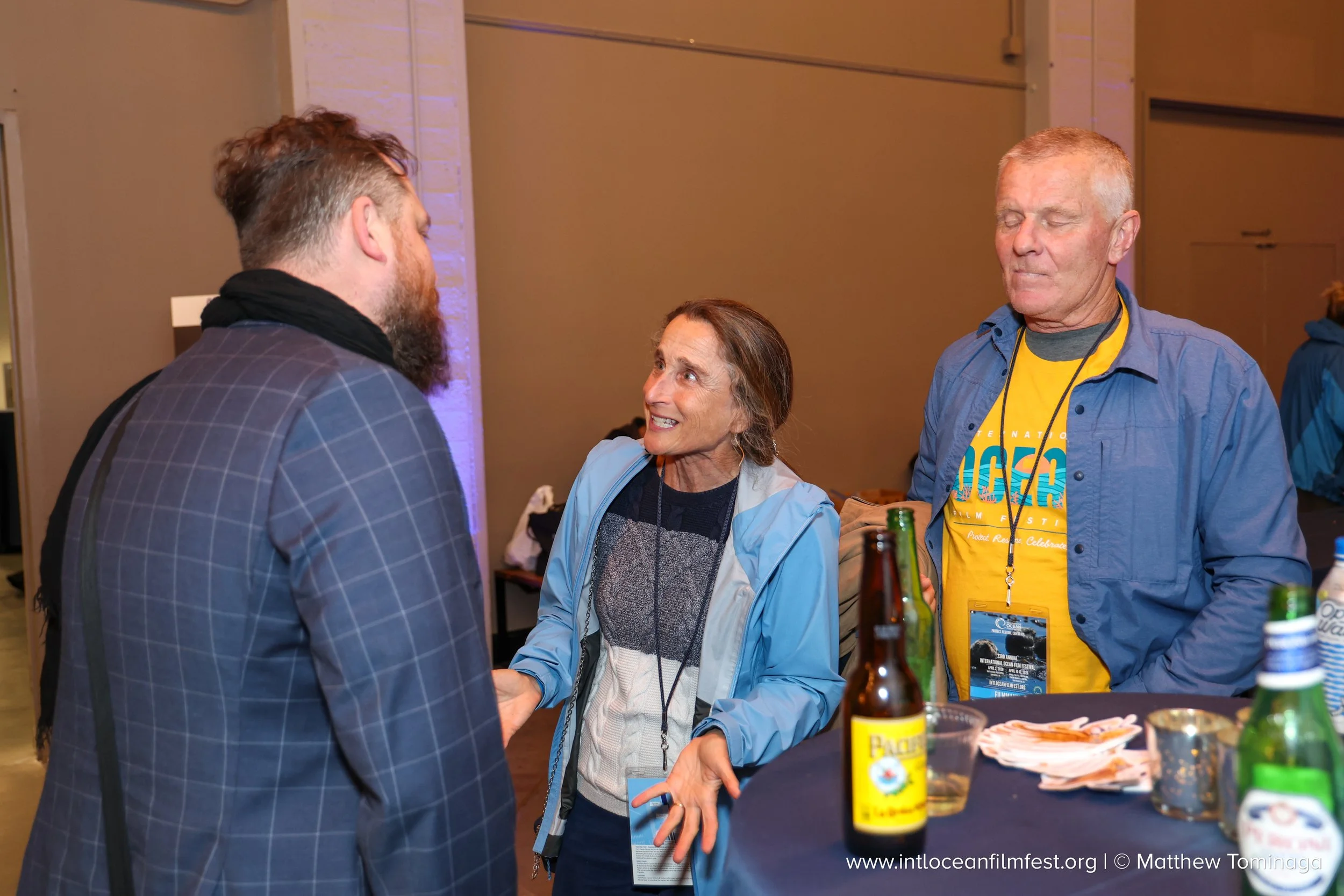 Three people are engaged in conversation at an indoor event. The woman in the center is smiling and talking to the two men on either side of her. They are standing near a table with bottles and snacks, and she is wearing a light blue jacket, while th