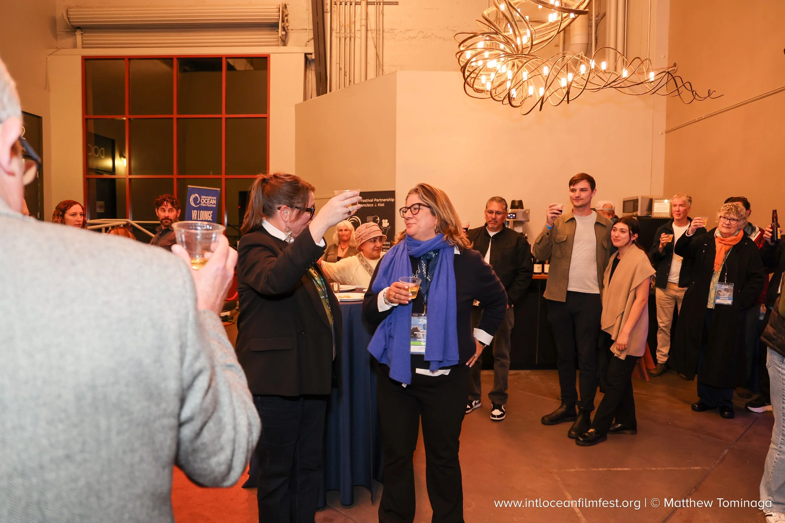A diverse group of people socializing at an indoor event with warm lighting, some holding drinks and engaging in conversation, and a large, decorative chandelier hanging from the ceiling.