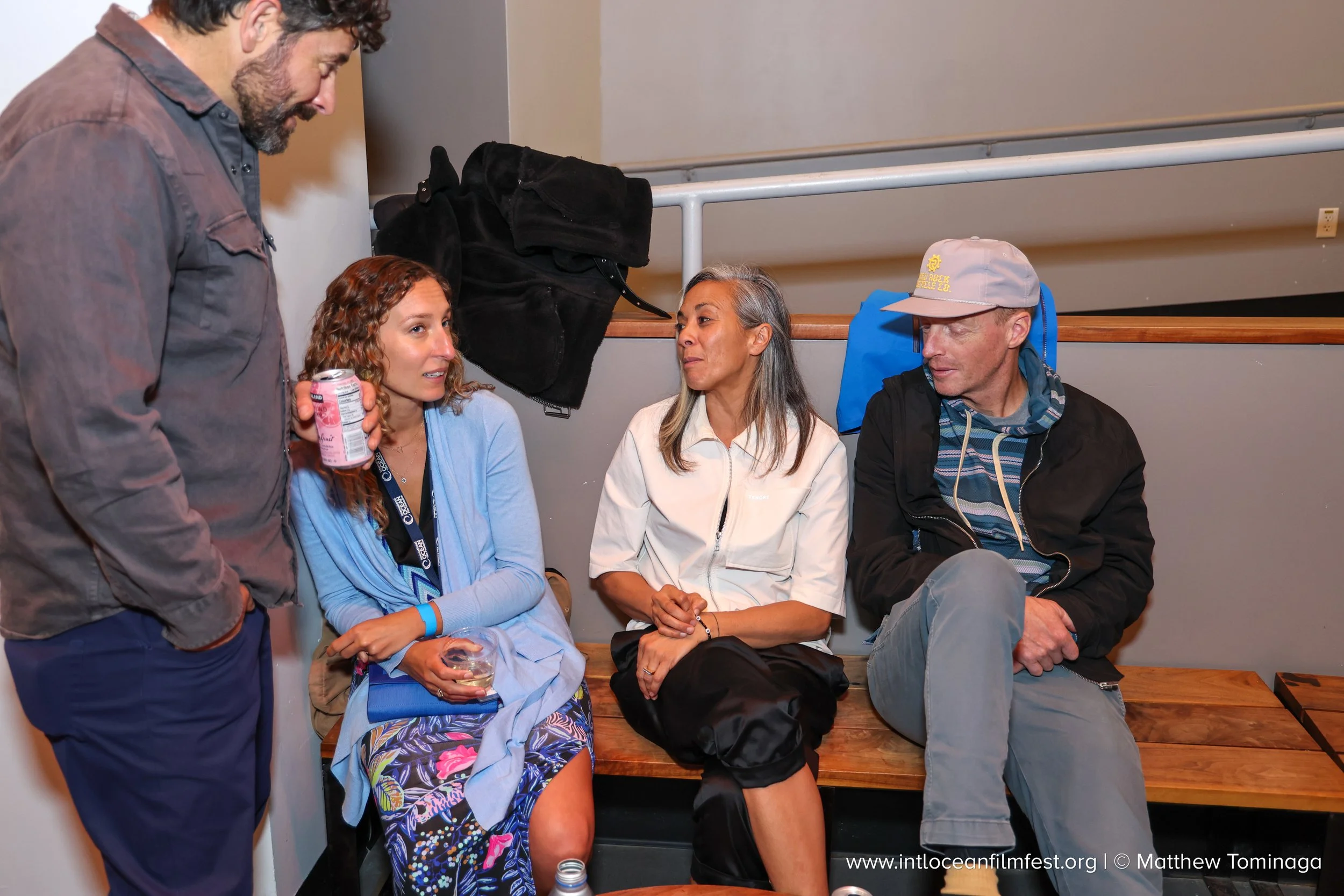 Four people are sitting on a wooden bench in a room, engaging in conversation. One man stands to the left holding a can, while three women sit, listening and talking. The women are dressed casually, and a black jacket hangs on a wall behind them. One
