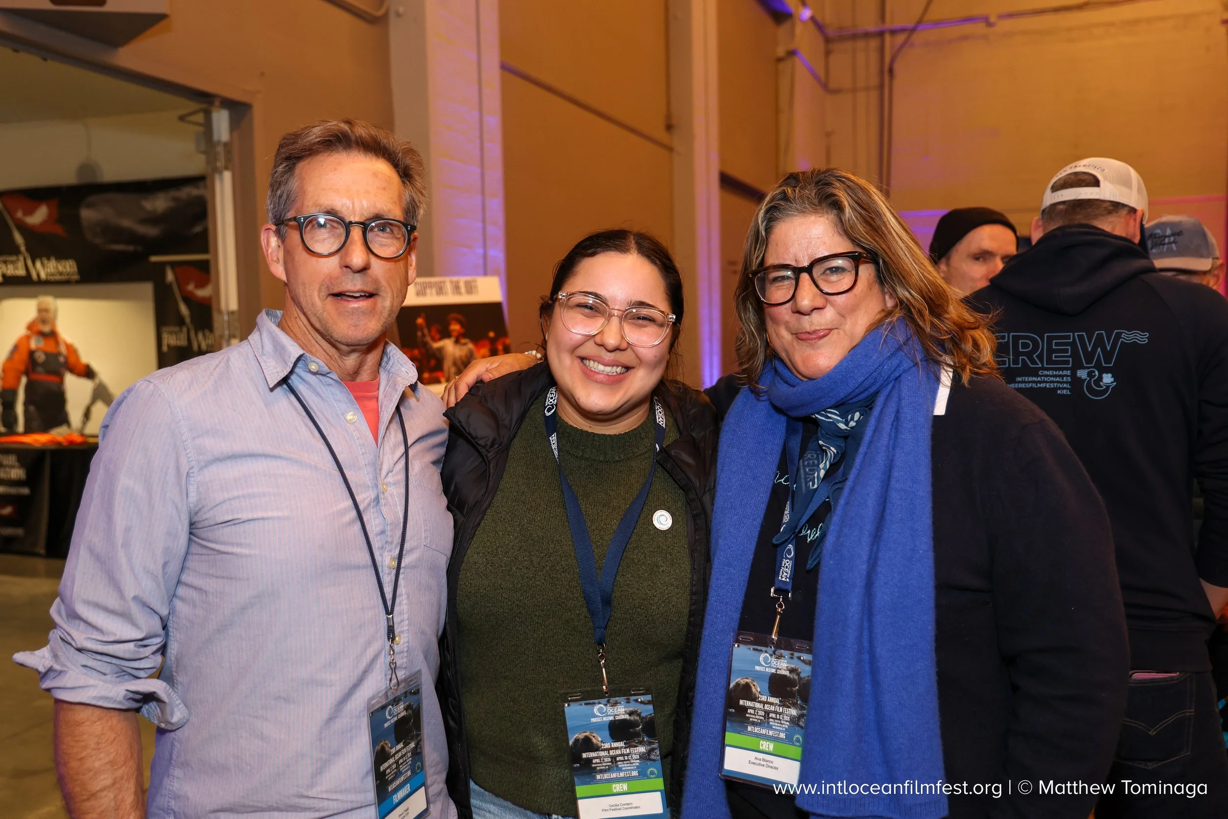 Three smiling people wearing conference badges are posing for a photo at the international ocean film festival, with others in the background at the event.