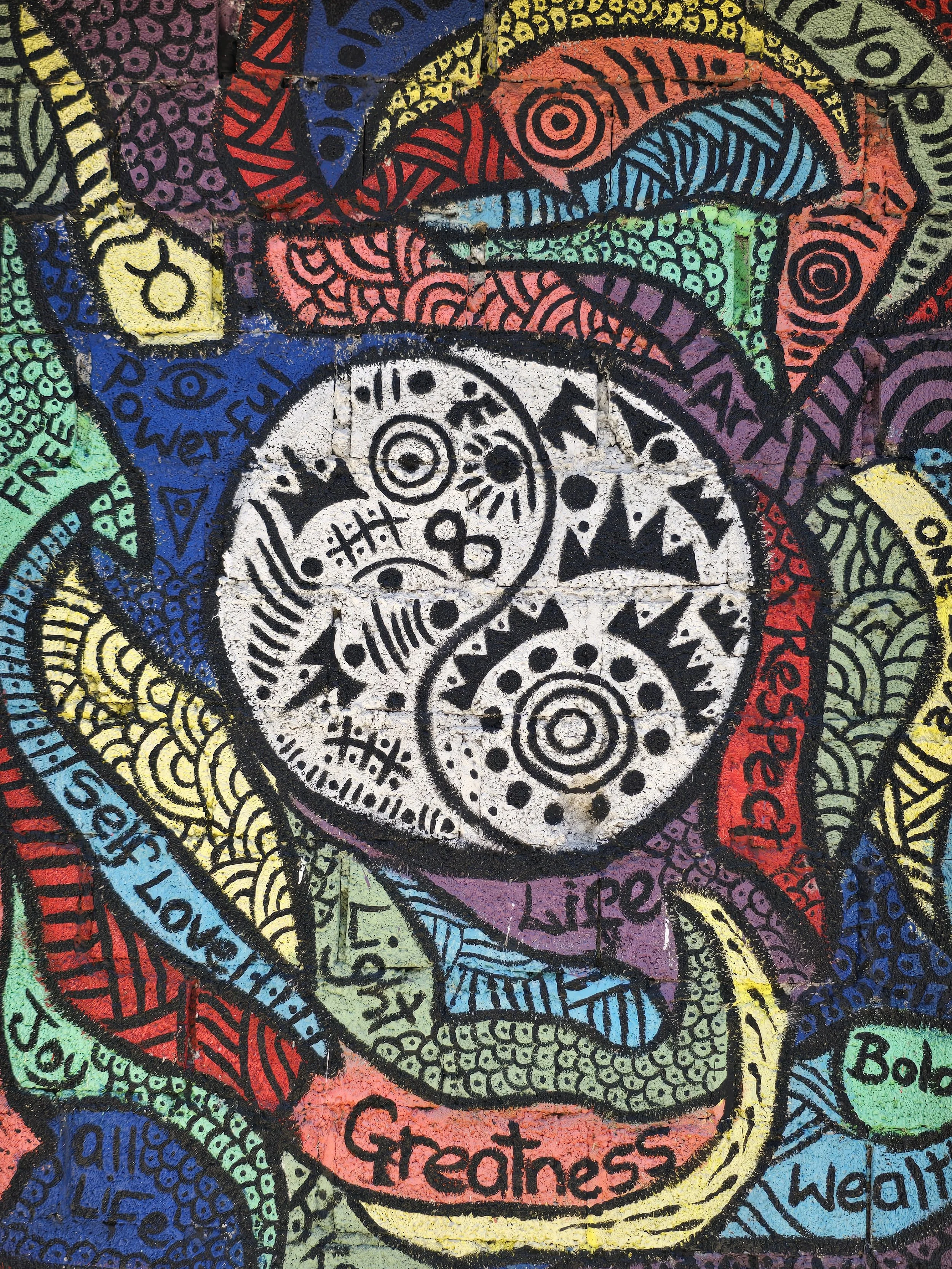 Colorful graffiti street art featuring a black and white yin-yang symbol with abstract designs surrounded by various words like 'Freedom,' 'Love,' 'Greatness,' and 'Wisdom' in different colors.