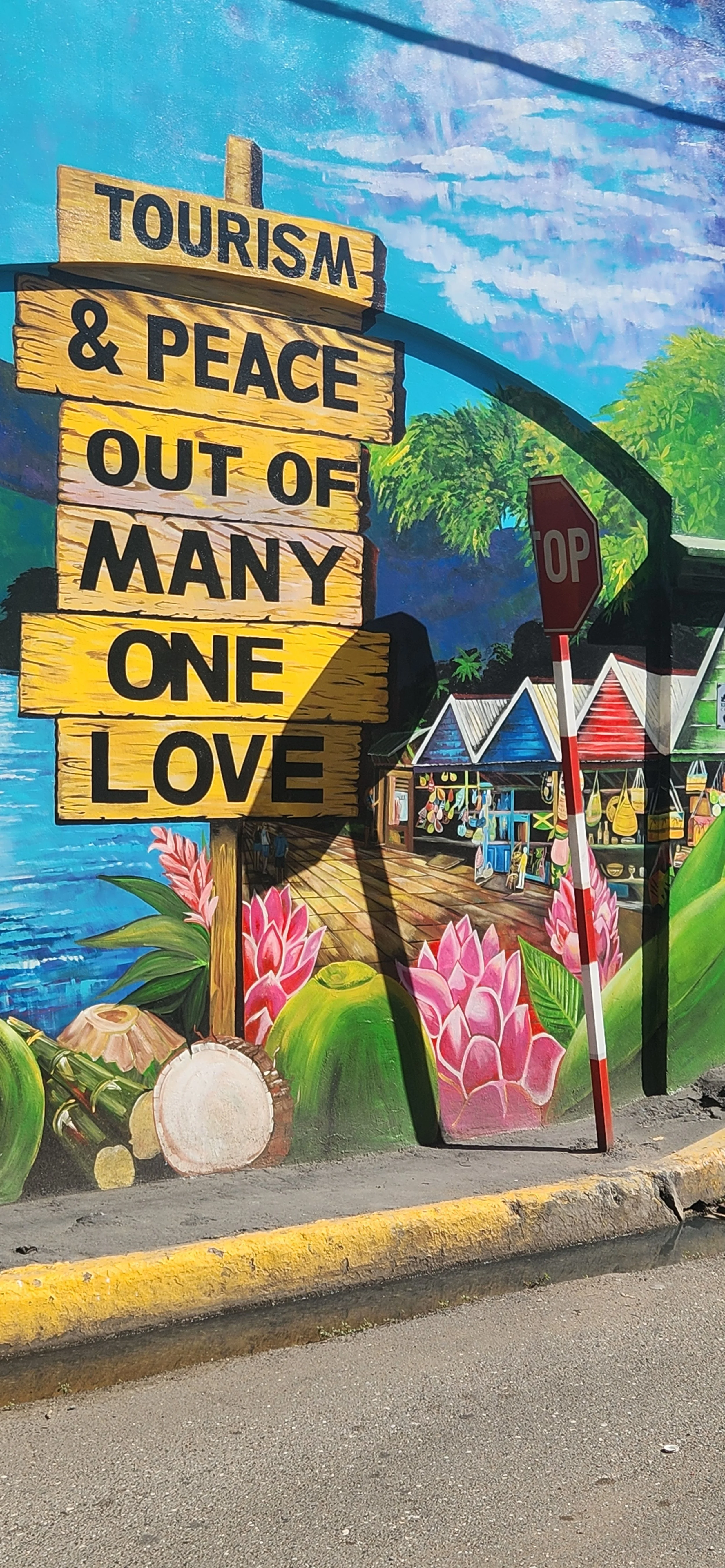 Colorful mural promoting tourism and peace, featuring a wooden sign with the text "Tourism & Peace Out of Many One Love", surrounded by vibrant flowers, tropical scenery, market stalls, a stop sign, and a bright blue sky.