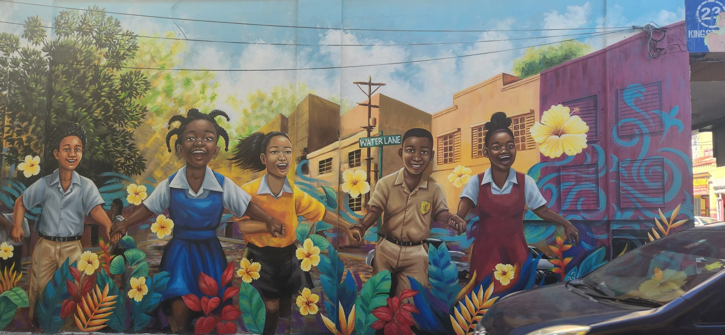 Colorful mural of six smiling children holding hands and playing, surrounded by flowers, with a city street and buildings in the background, including a street sign for Water Lane.