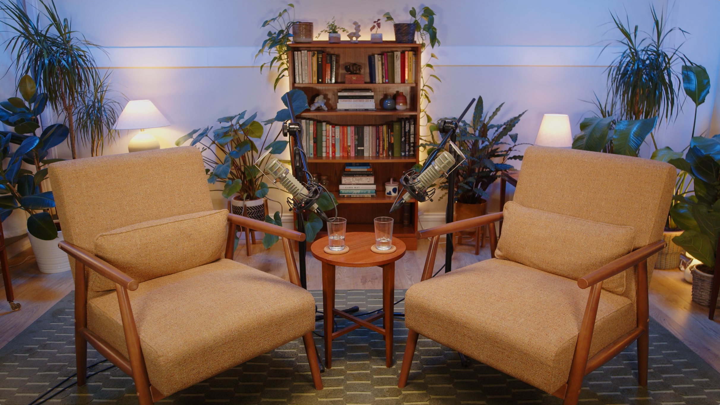 A cozy living room setup for a podcast recording with two beige armchairs facing each other, microphones on stands, a small round wooden table with two glasses, potted plants, a bookshelf with books, and warm lighting from lamps.